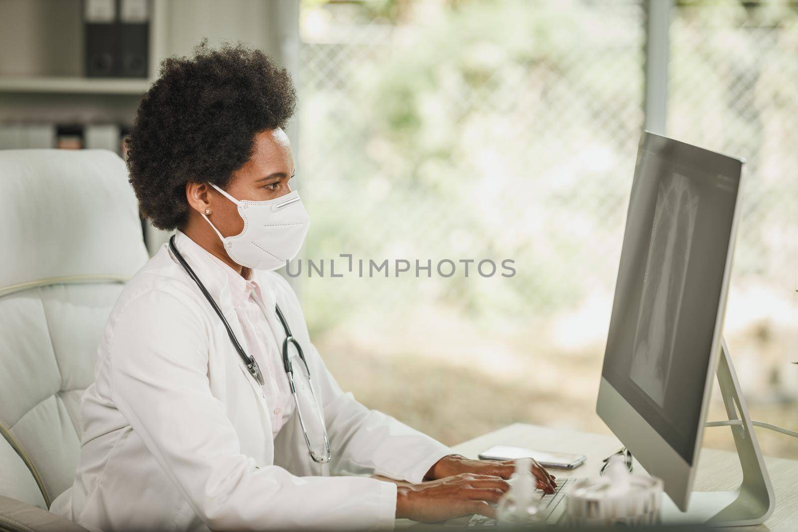 Shot of an African female doctor sitting alone in her consulting room and looking X-ray on computer during COVID-19 pandemic.