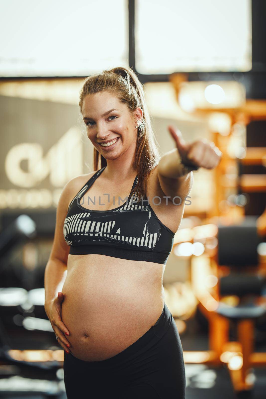 Shot of a pregnant woman resting from her workout at the gym. She is holding her pregnant belly and showing OK sign with her thumb up.