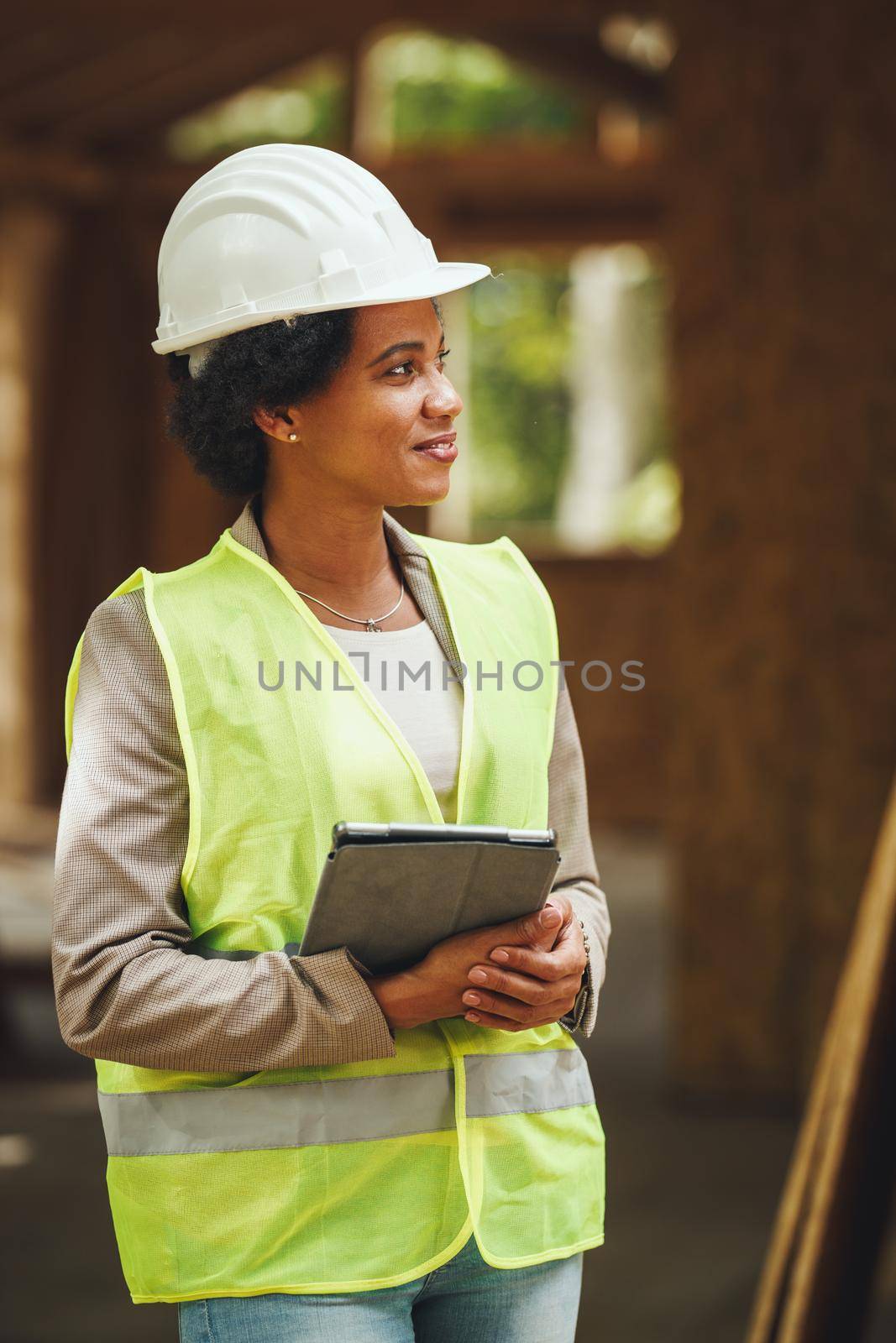 Shot of an African female architect using a digital tablet and checking construction site of a new wooden house. She is wearing protective workwear and white helmet.