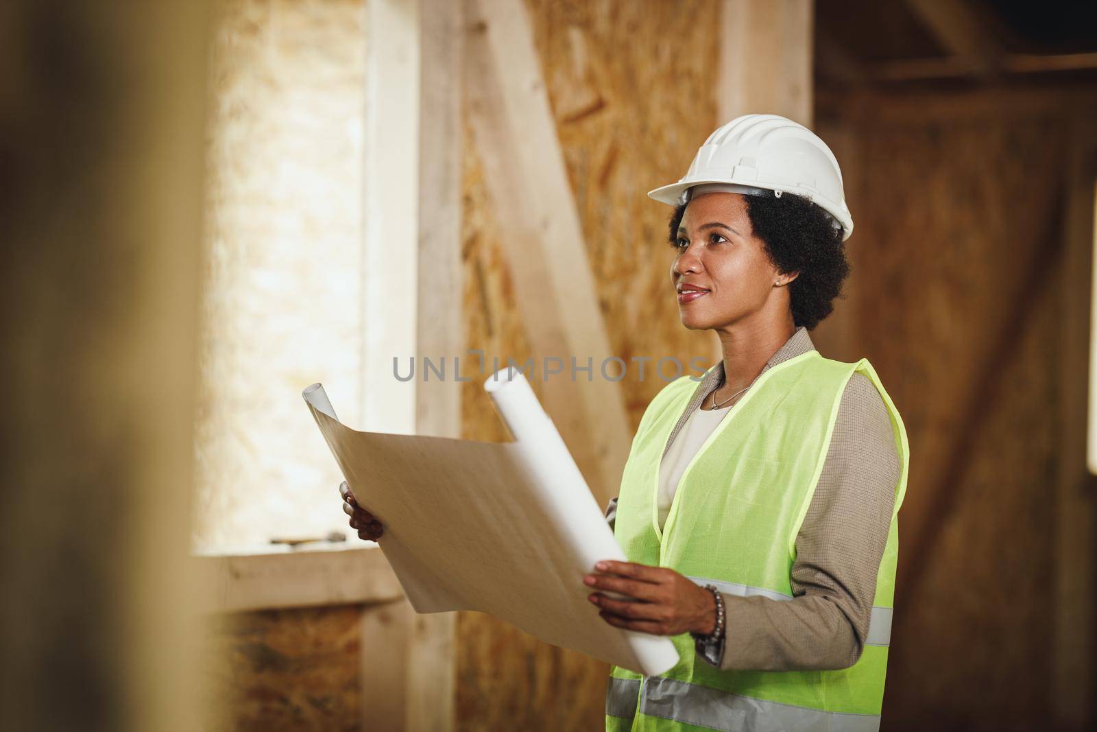 Shot of an African female architect checking blueprints at the construction site of a new wooden house. She is wearing protective workwear and white helmet.