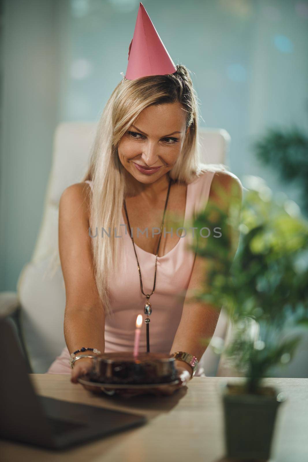 Smiling attractive woman have birthday celebration at home during pandemic isolation and have video call with friends. She holding birthday cake with lighted candles.