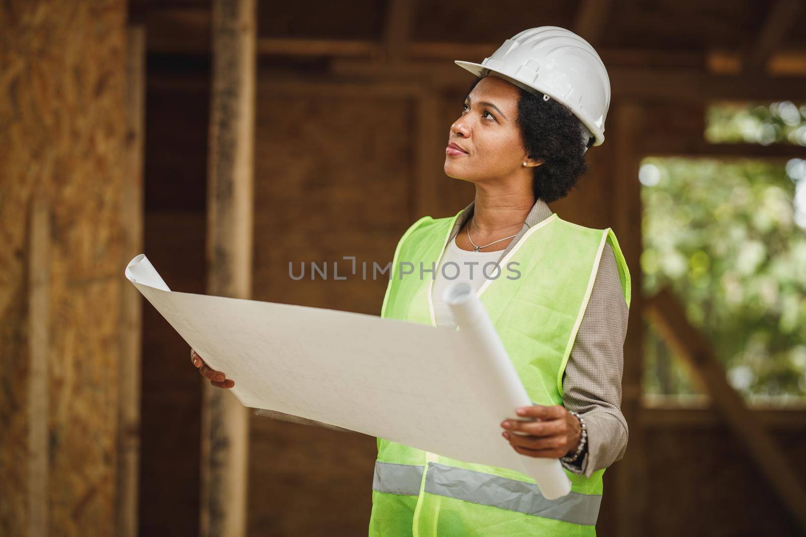 Shot of an African female architect checking blueprints at the construction site of a new wooden house. She is wearing protective workwear and white helmet.