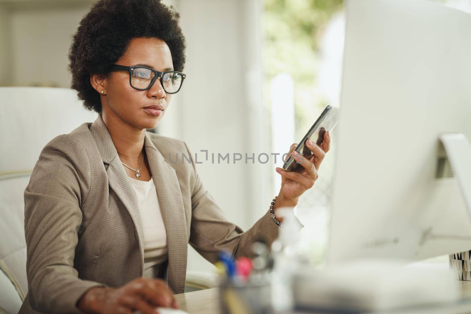 Shot of an African businesswoman using smartphone while working at computer in her home office during COVID-19 pandemic.