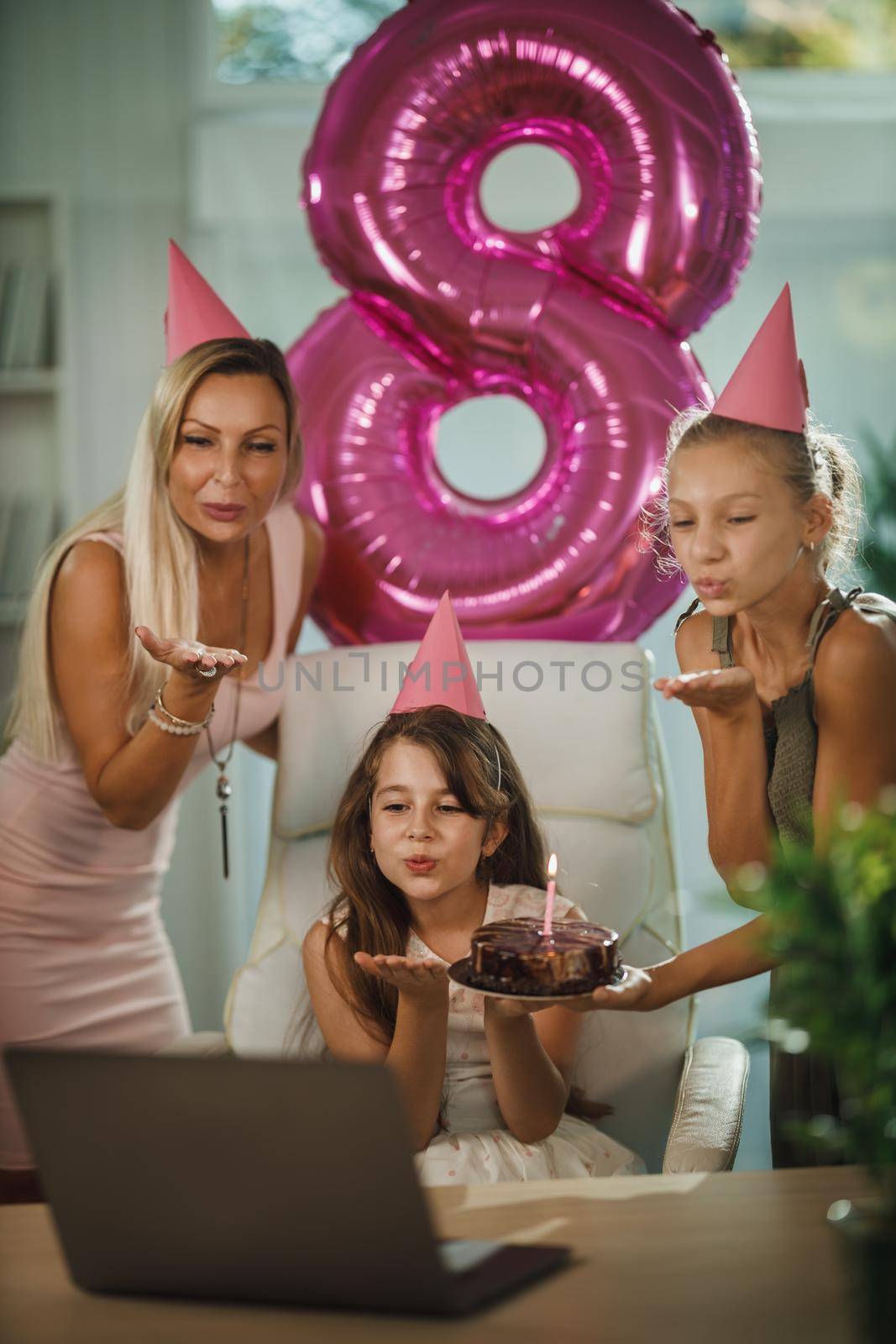 Beautiful young mom with her daugters at home during pandemic isolation have birthday party, she blowing candles and have conference call with friends and family.