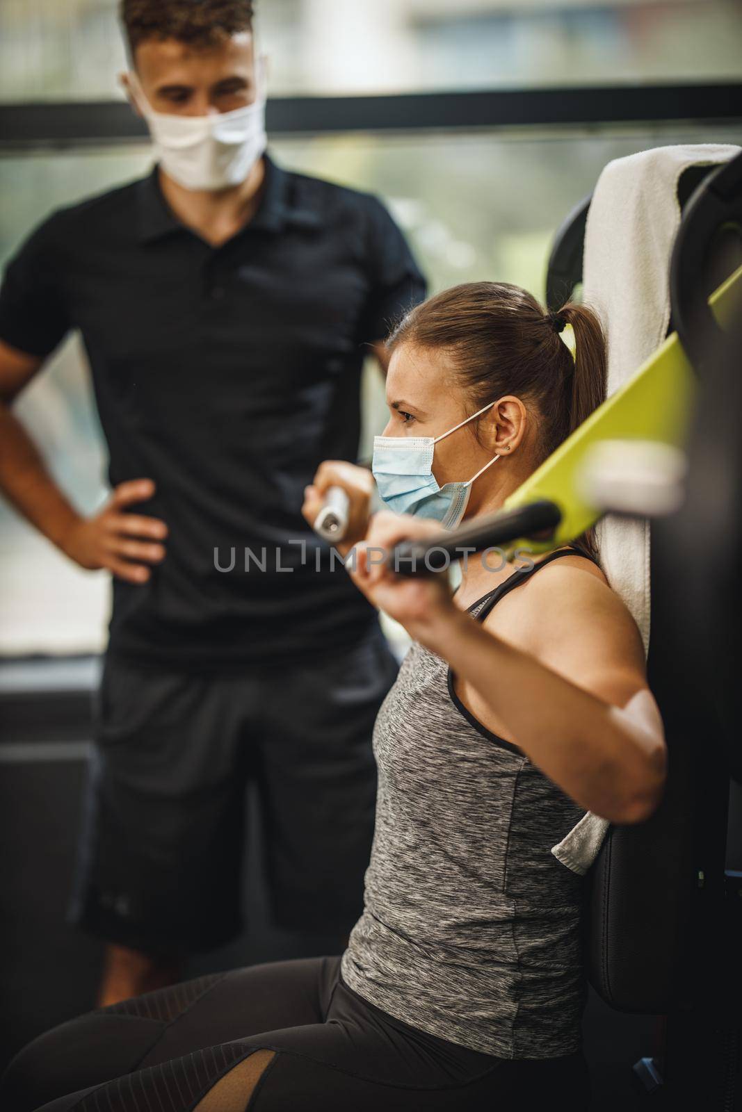 Shot of a muscular young woman with protective mask working out with personal trainer at the gym machine during Covid-19 pandemic. She is pumping up her shoulder muscule with heavy weight.