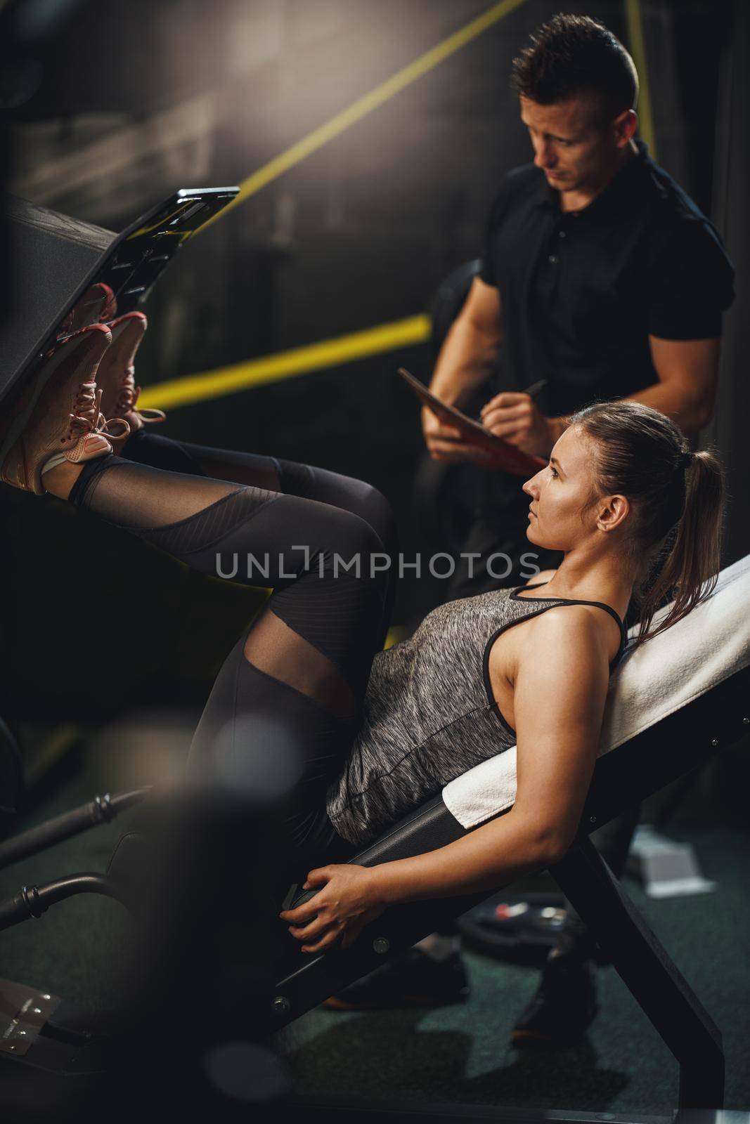 Shot of a muscular young woman in sportswear working out with personal trainer at the gym. She is doing exercises for her legs on leg press machine.