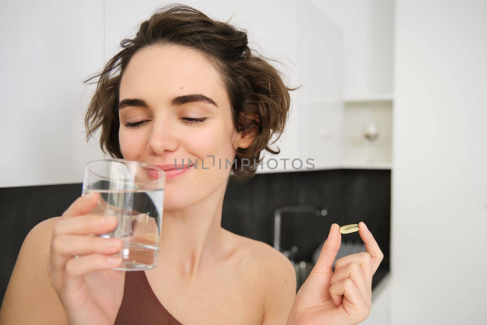 Portrait of sportswoman drinking water, taking vitamins, dietry supplements for healthy skin, having omega-3 pill, standing in her kitchen in workout clothing.