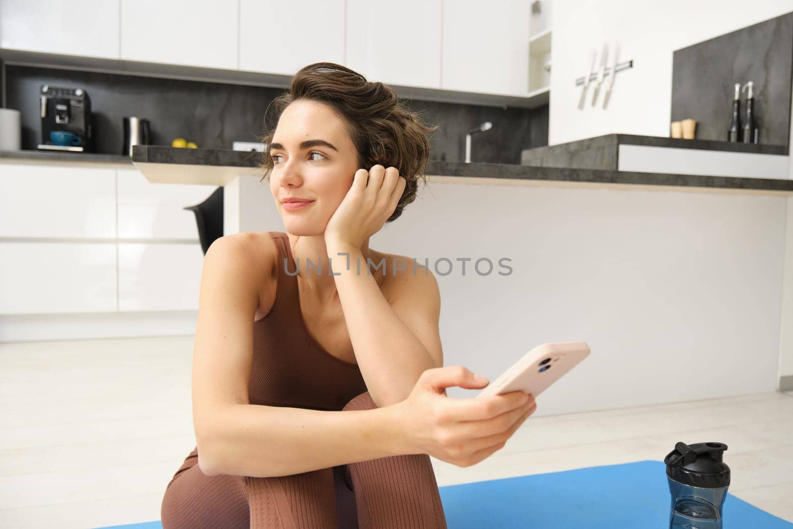 Portrait of sporty girl with mobile phone, sitting on yoga mat, using smartphone during workout at home, watching fitness turorial on telephone by Benzoix