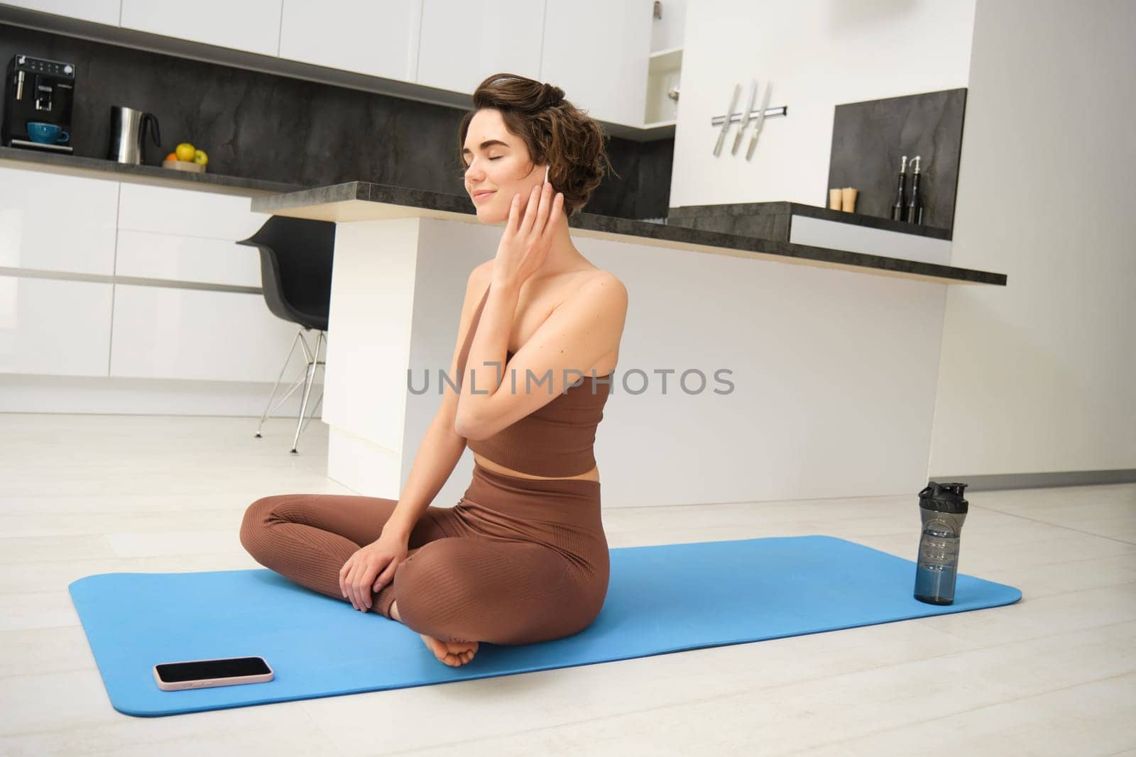 Mindfulness and healthy lifestyle. Young smiling woman sits on floor at home, practice yoga in earphones, listens to meditation app on smartphone, uses mat for exercises and workout by Benzoix