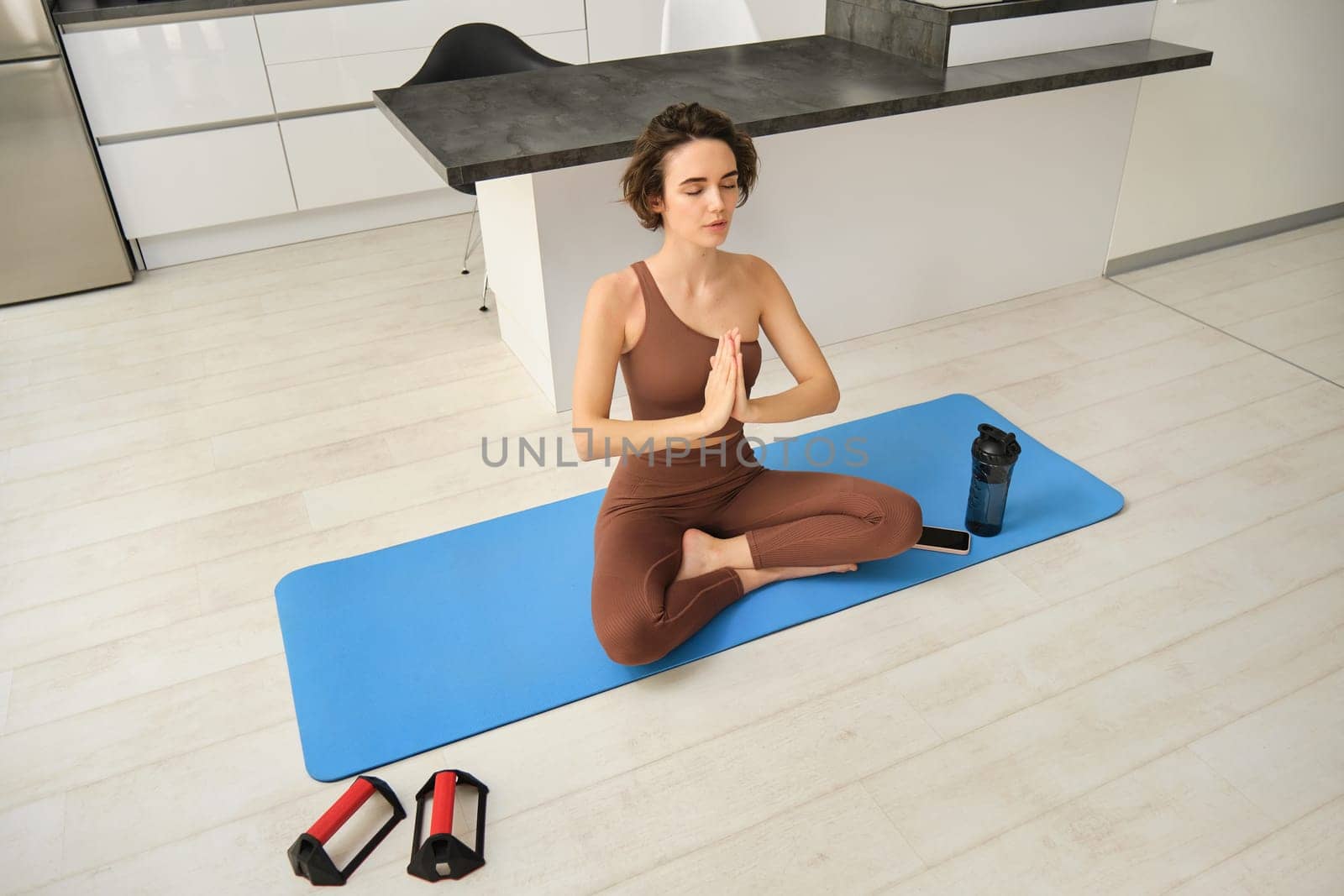 Young calm fit healthy woman wearing sportswear sitting at home in living room doing yoga exercise, meditating and breathing in the morning. Mental health and zen meditation concept.