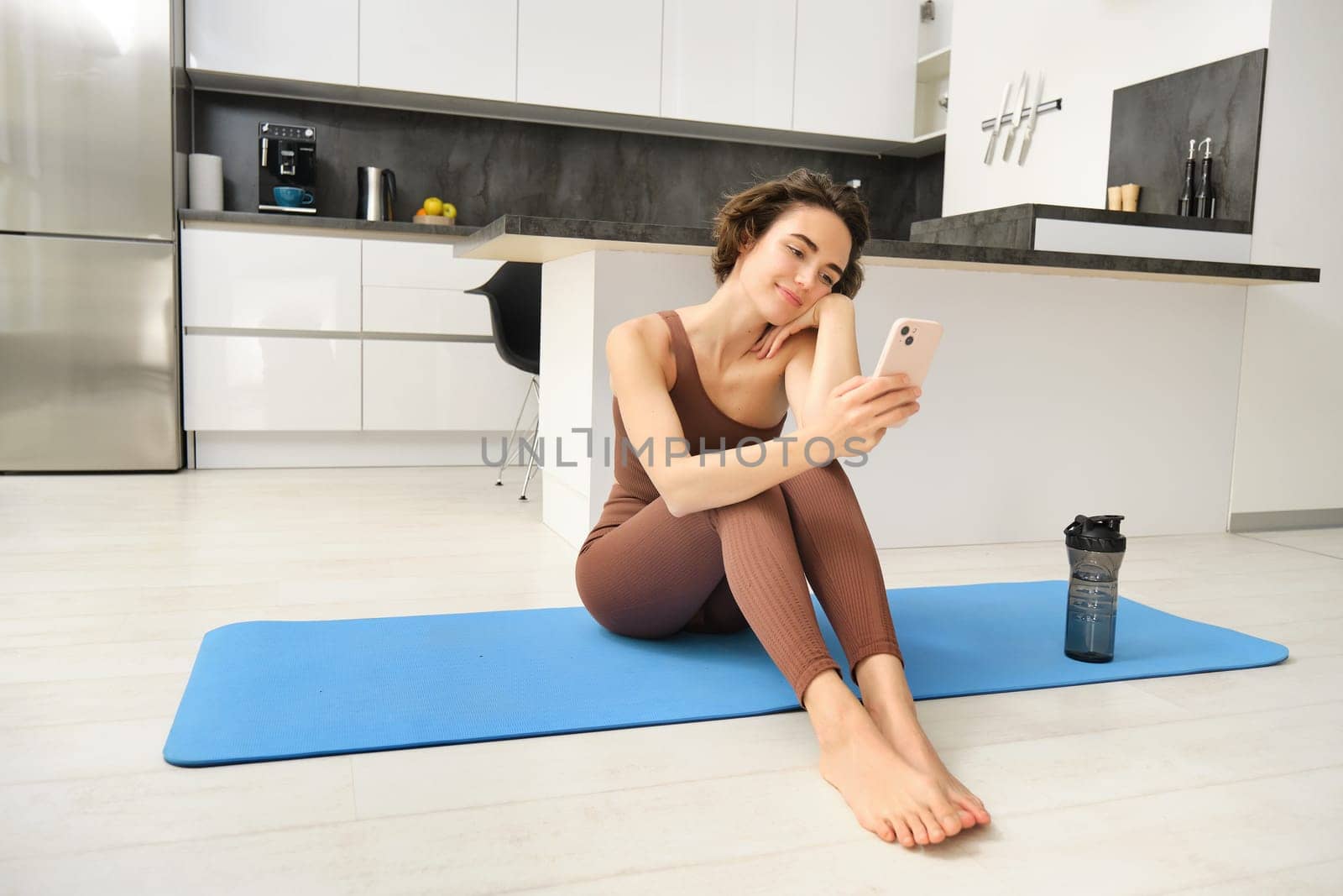 Sport and wellbeing. Portrait of fitness woman, gym instructor workout at home, sits on yoga mat and looks at her smartphone with smiling face by Benzoix