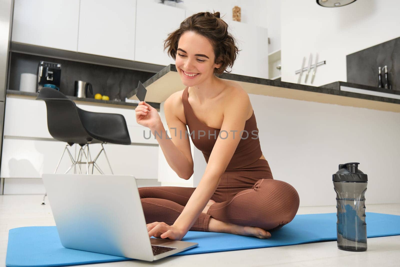 Image of young fitness girl, female athlete at home, workout on kitchen floor, holding credit card with laptop, purchasing online training session, gym application for home workout by Benzoix