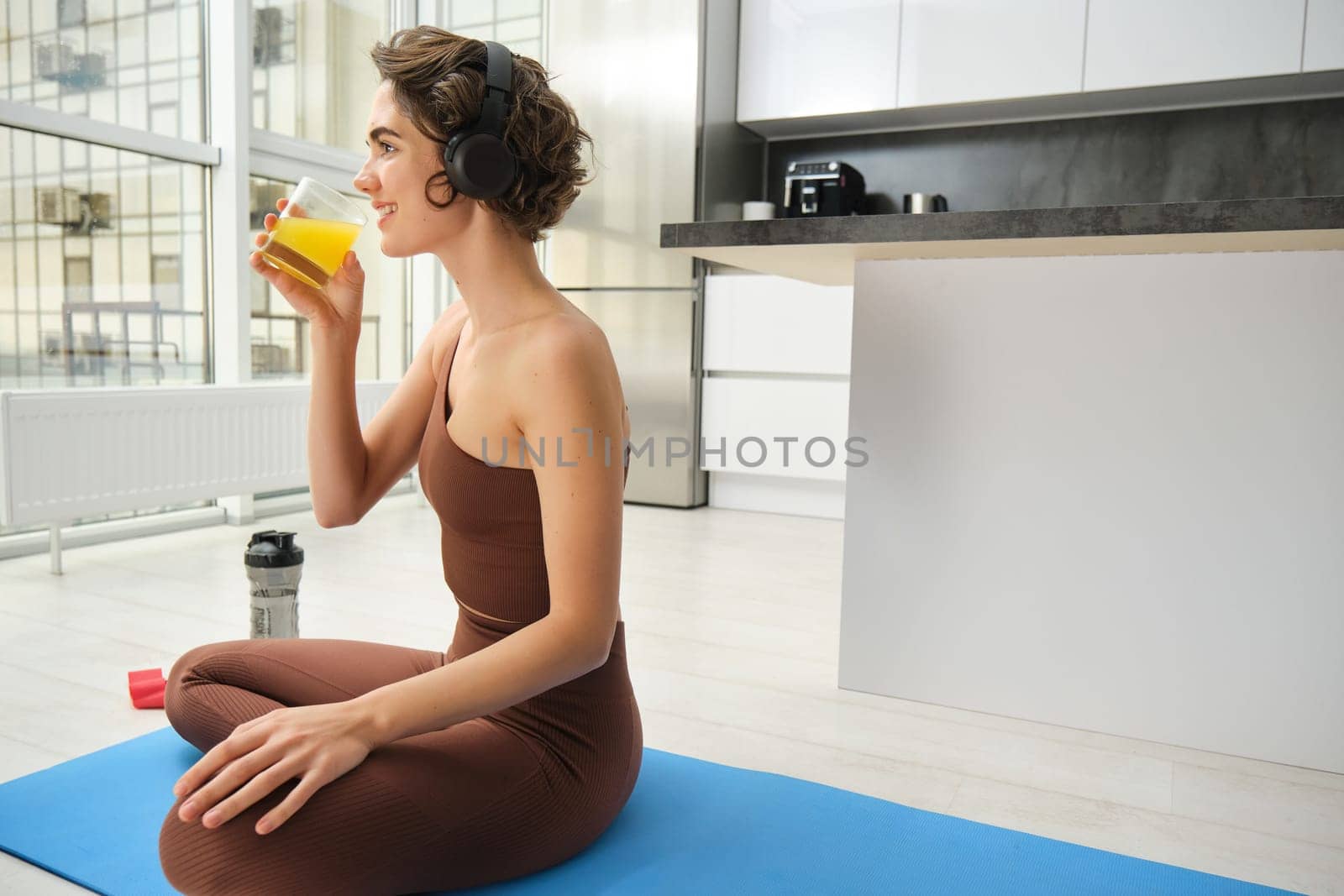 Sport and healthy lifestyle. Sportswoman at home, workout, drinks orange juice after fitness training, doing sports indoors on rubber mat by Benzoix