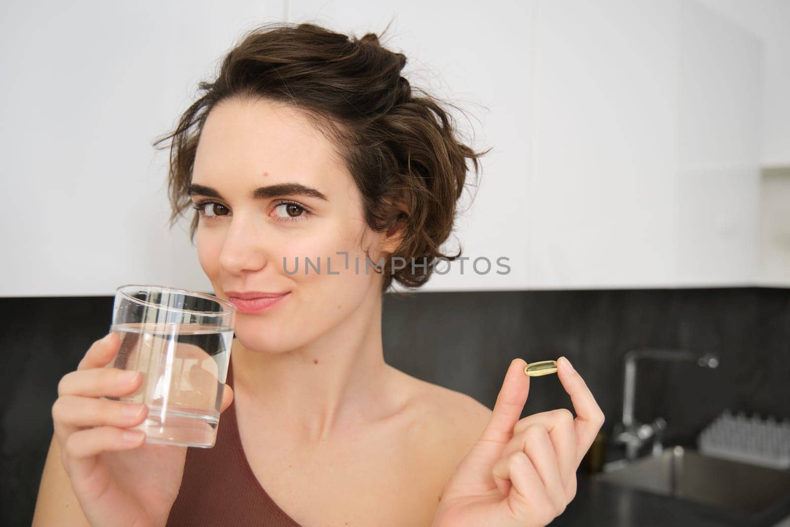 Dietry supplements and healthy lifestyle. Young woman taking vitamin C, D omega-3 with glass of water, standing in activewear, drinking after workout training in her kitchen by Benzoix