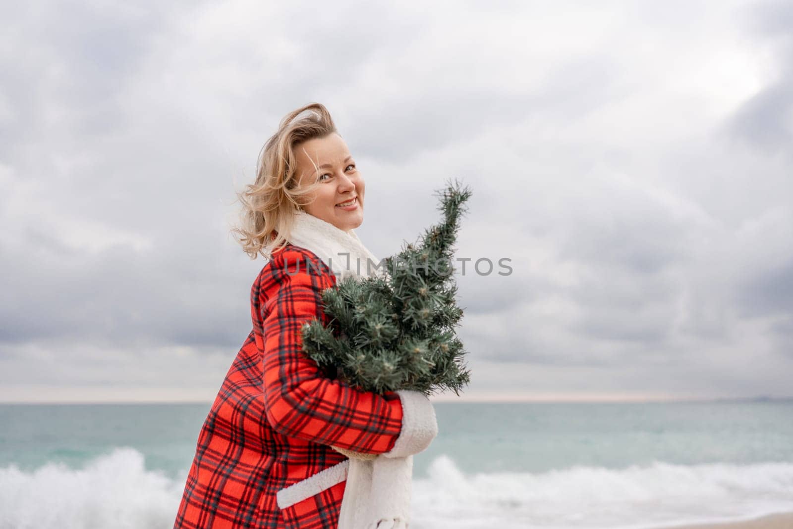 Blond woman holding Christmas tree by the sea. Christmas portrait of a happy woman walking along the beach and holding a Christmas tree in her hands. Dressed in a red coat, white dress. by Matiunina