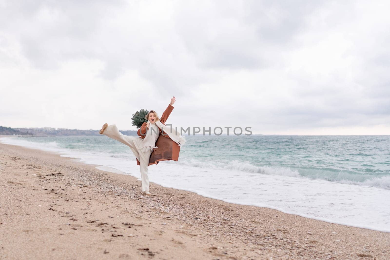 Blond woman Christmas sea. Christmas portrait of a happy woman walking along the beach and holding a Christmas tree on her shoulder. She is wearing a brown coat and a white suit