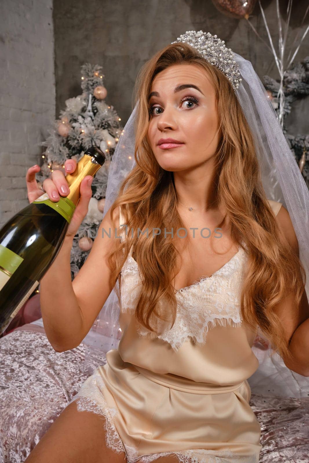 Gorgeous blonde lady in lacy beige lingerie, white veil and tiara is enjoying hen-party of bride, sitting on bed and drinking champagne. Christmas tree, decorations, balloons. Fashion. Close-up.