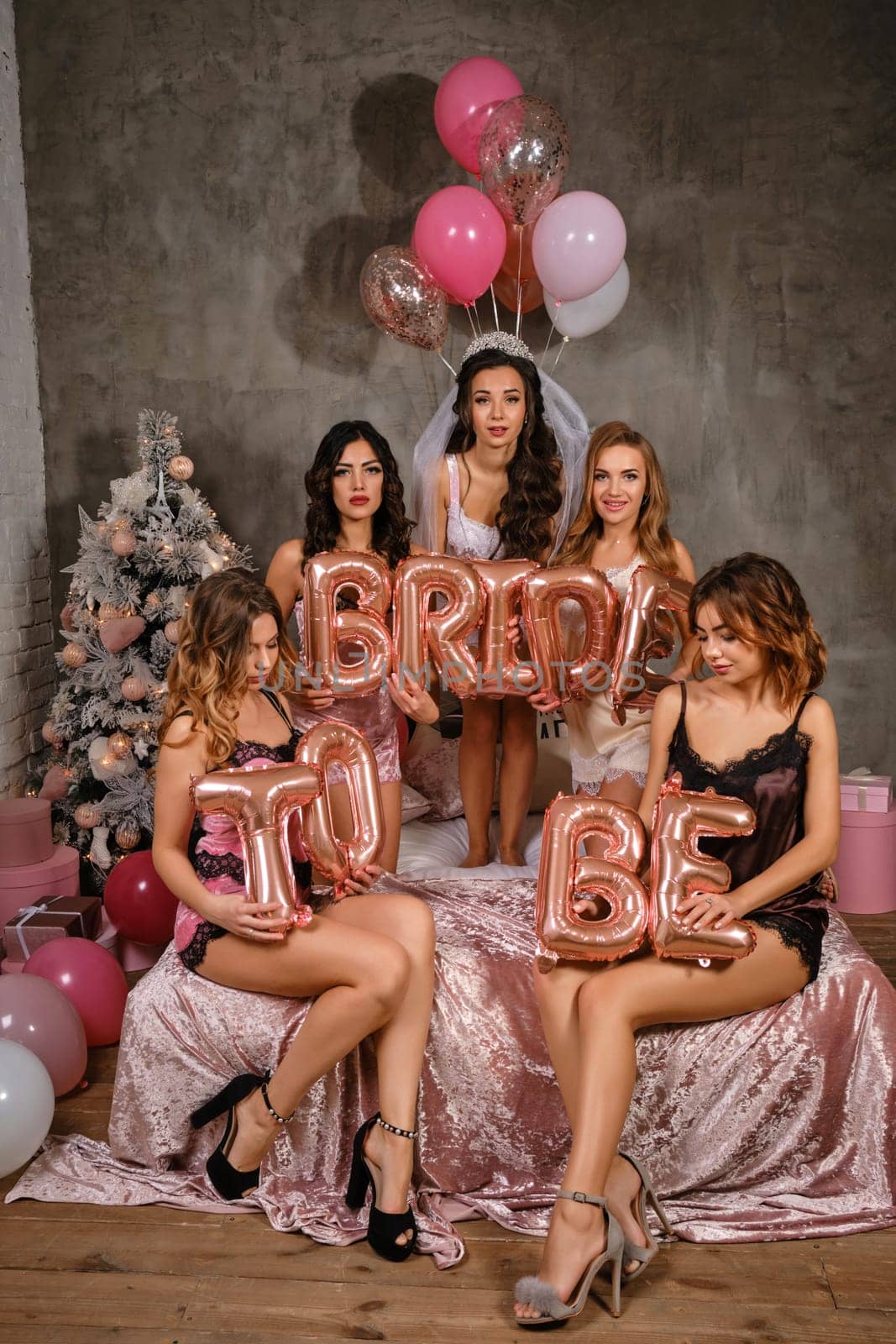 Cute females in sexy lingerie, bride in veil, enjoying bachelorette party, sitting on bed, smiling, holding balloons in form of letters, built sentence bride to be. Christmas decorations. Close-up.