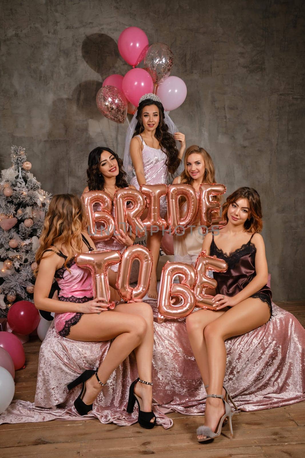 Charming maidens in sexy lingerie, bride in veil, enjoying hen-party, sitting on bed, smiling, holding balloons in form of letters, built sentence bride to be. Christmas tree, decorations. Close-up.