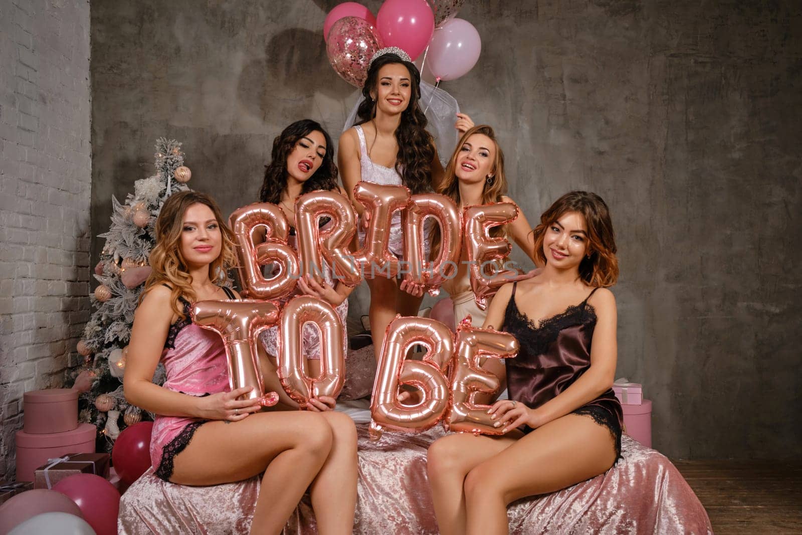 Pretty women in sexy lingerie and bride in veil, enjoying bachelorette party, sitting on bed, smiling, holding balloons in form of letters, built sentence bride to be. Christmas decorations. Close-up.