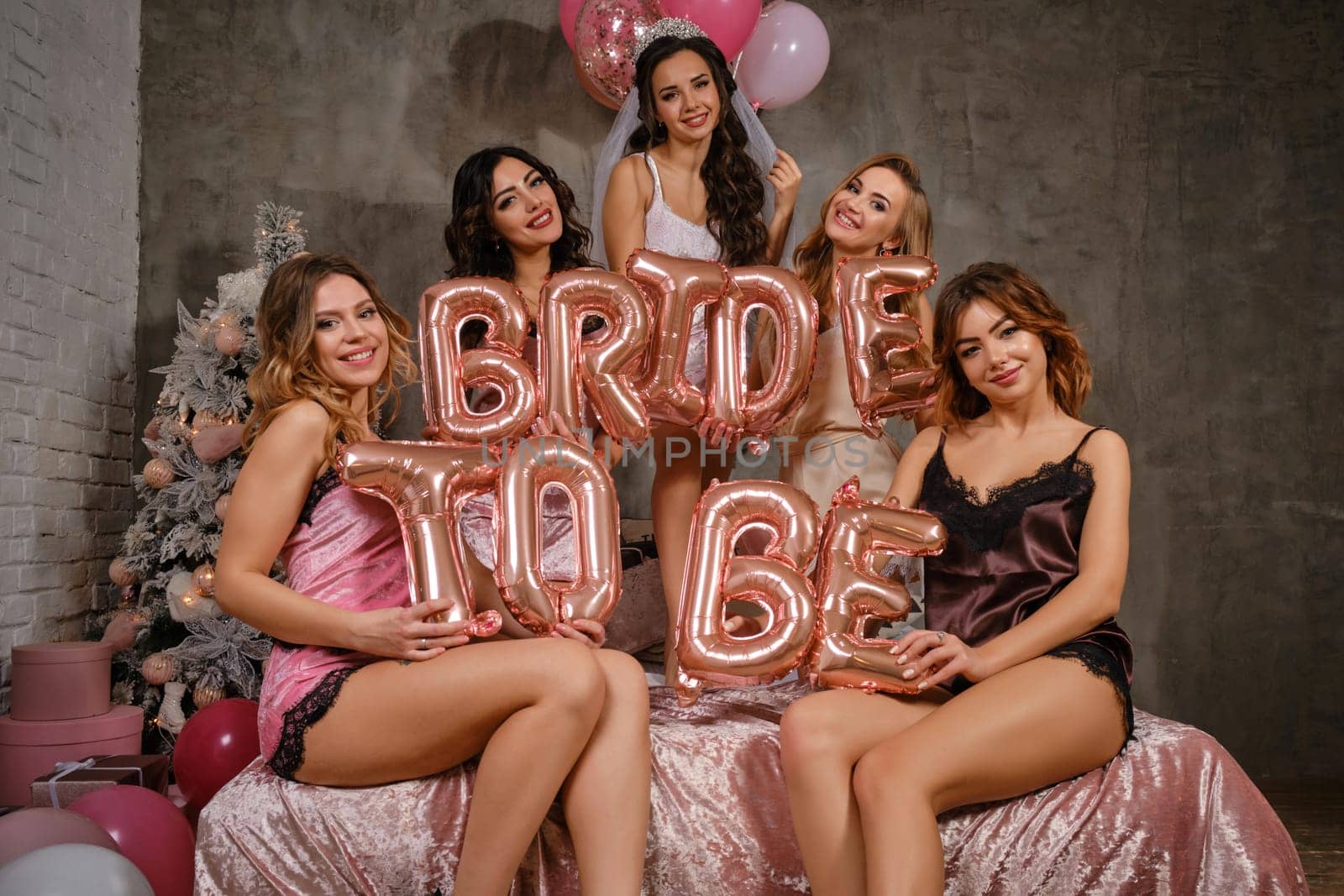 Pretty females in sexy lingerie and bride in veil, enjoying hen-party, sitting on bed, smiling, holding balloons in form of letters, built sentence bride to be. Christmas tree, decorations. Close-up.