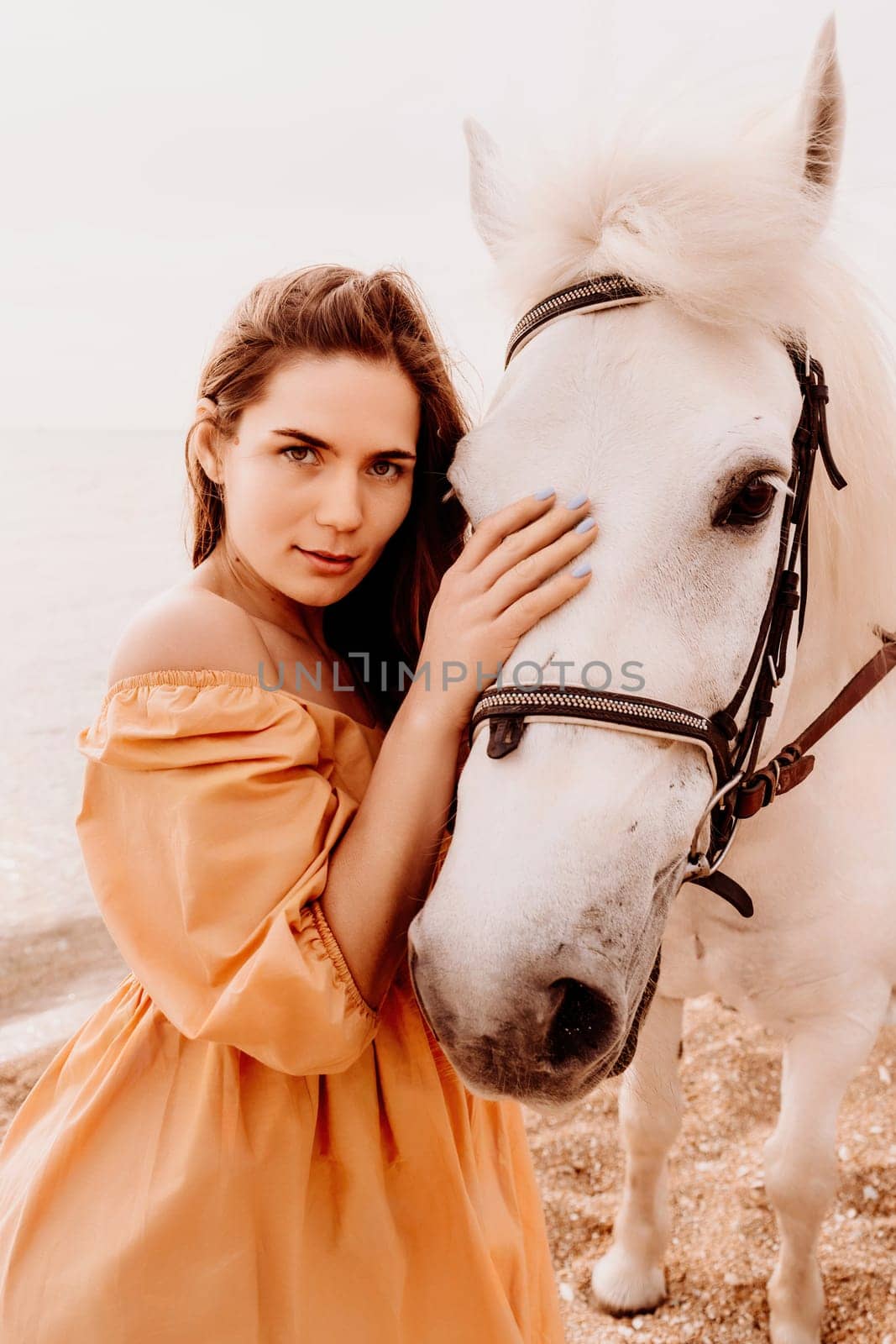 A white horse and a woman in a dress stand on a beach, with the sky and sea creating a picturesque backdrop for the scene