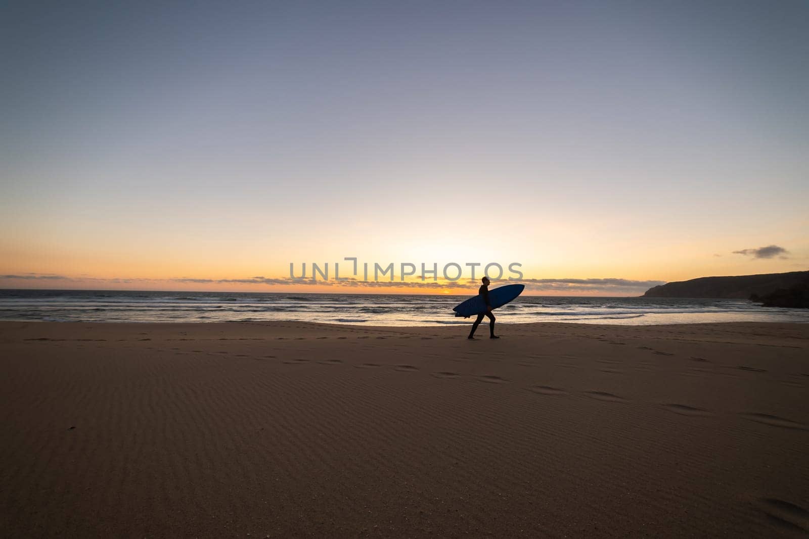Silhouette of an adult man walking on the empty beach holding a surfboard. Mid shot