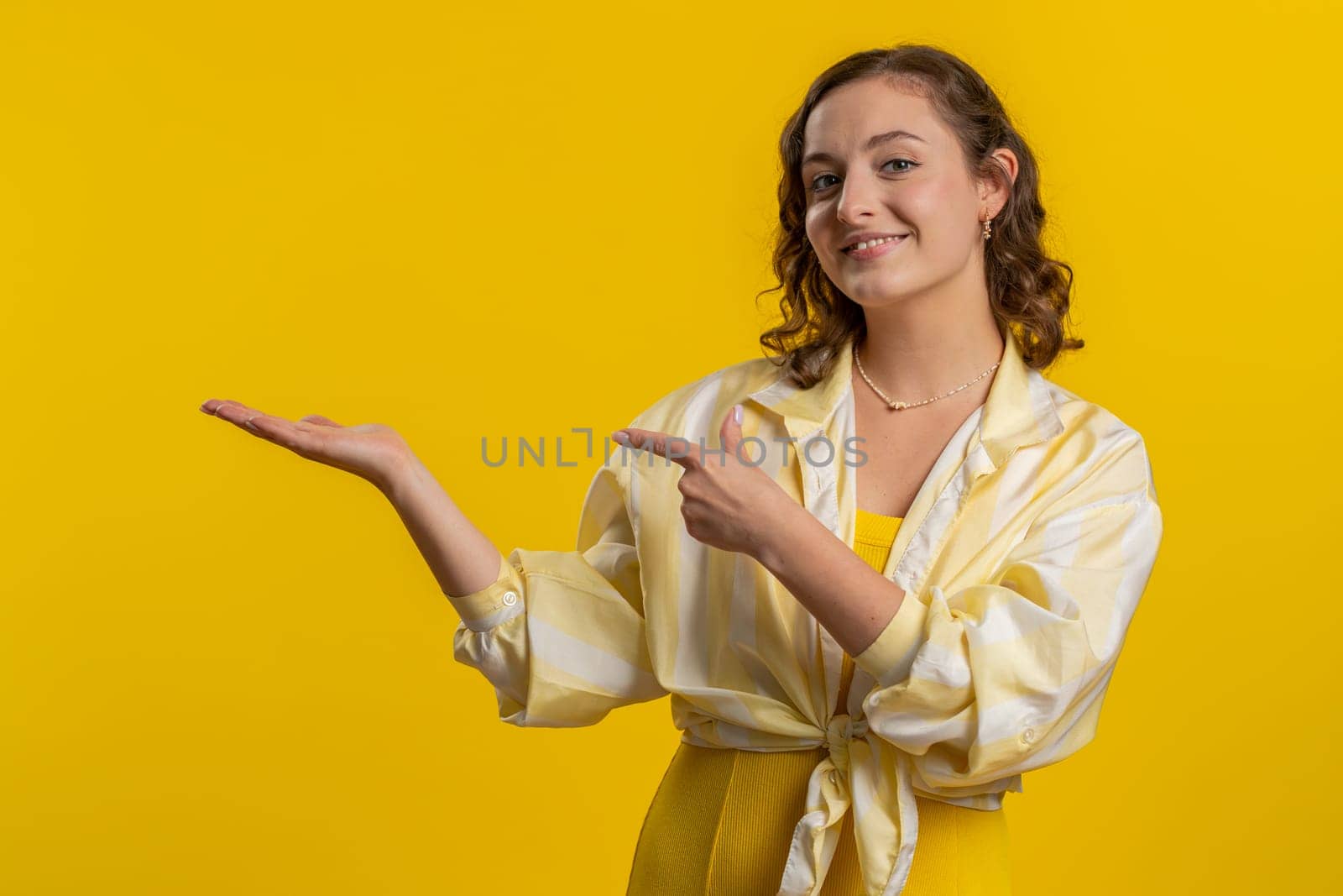 Young woman showing thumbs up and pointing empty place, advertising area for commercial text, copy space for goods promotion advertisement. Girl isolated alone on yellow studio background. Lifestyles