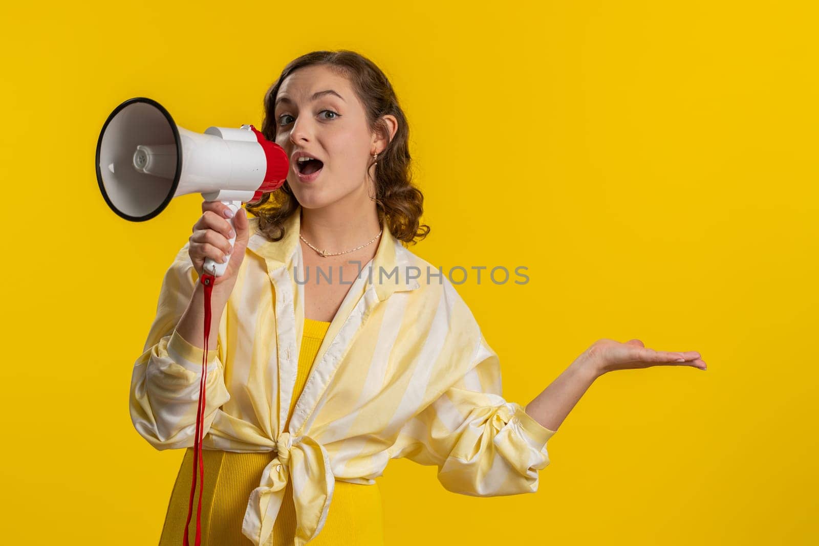 Woman talking with megaphone, proclaiming news, loudly announcing advertisement, warning using loudspeaker to shout speech, pointing empty place. Copy-space. Pretty brunette girl on yellow background