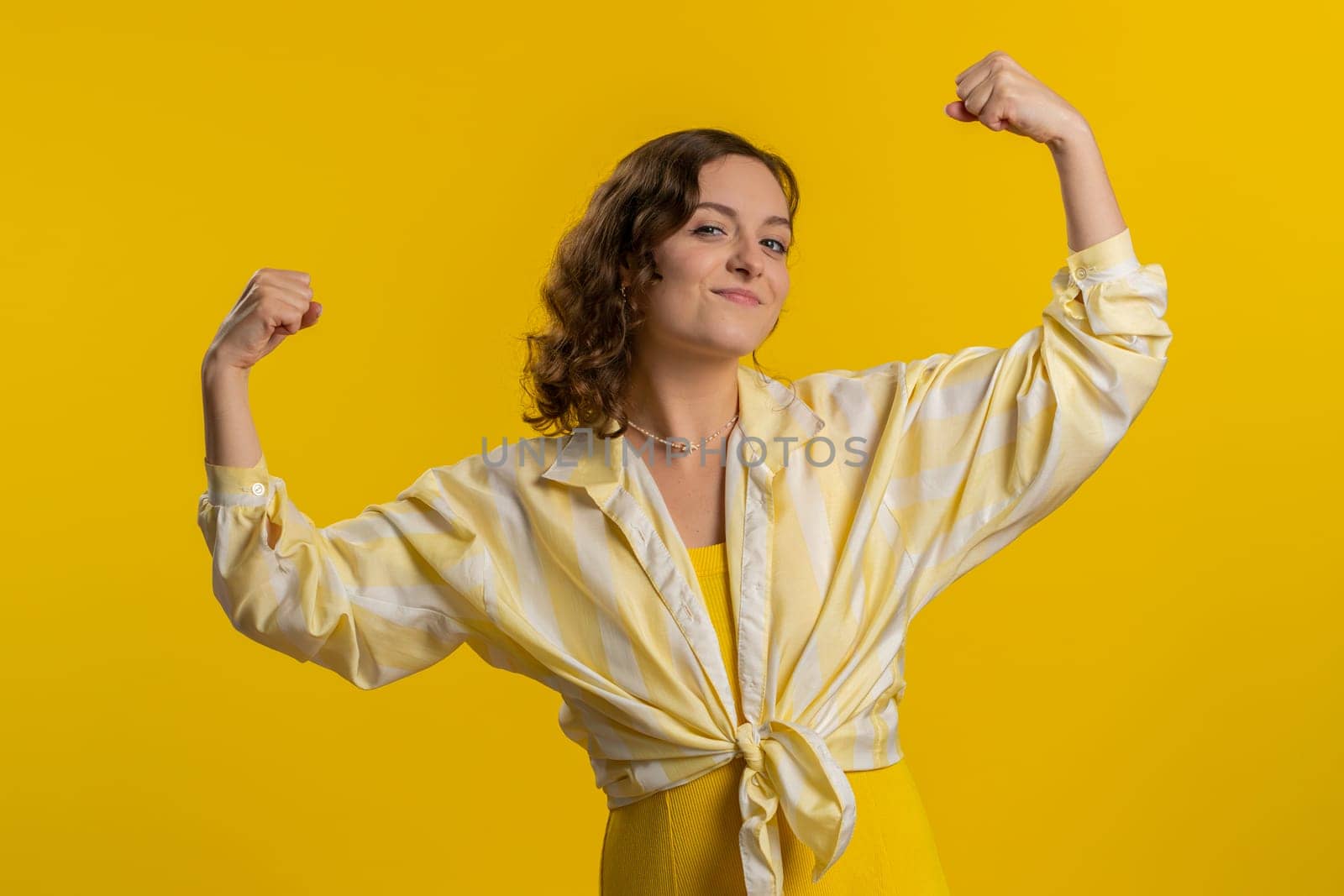 I am strong and independent. Young woman showing biceps and looking confident, feeling power strength to fight for rights, energy to gain success win. Girl isolated alone on yellow studio background