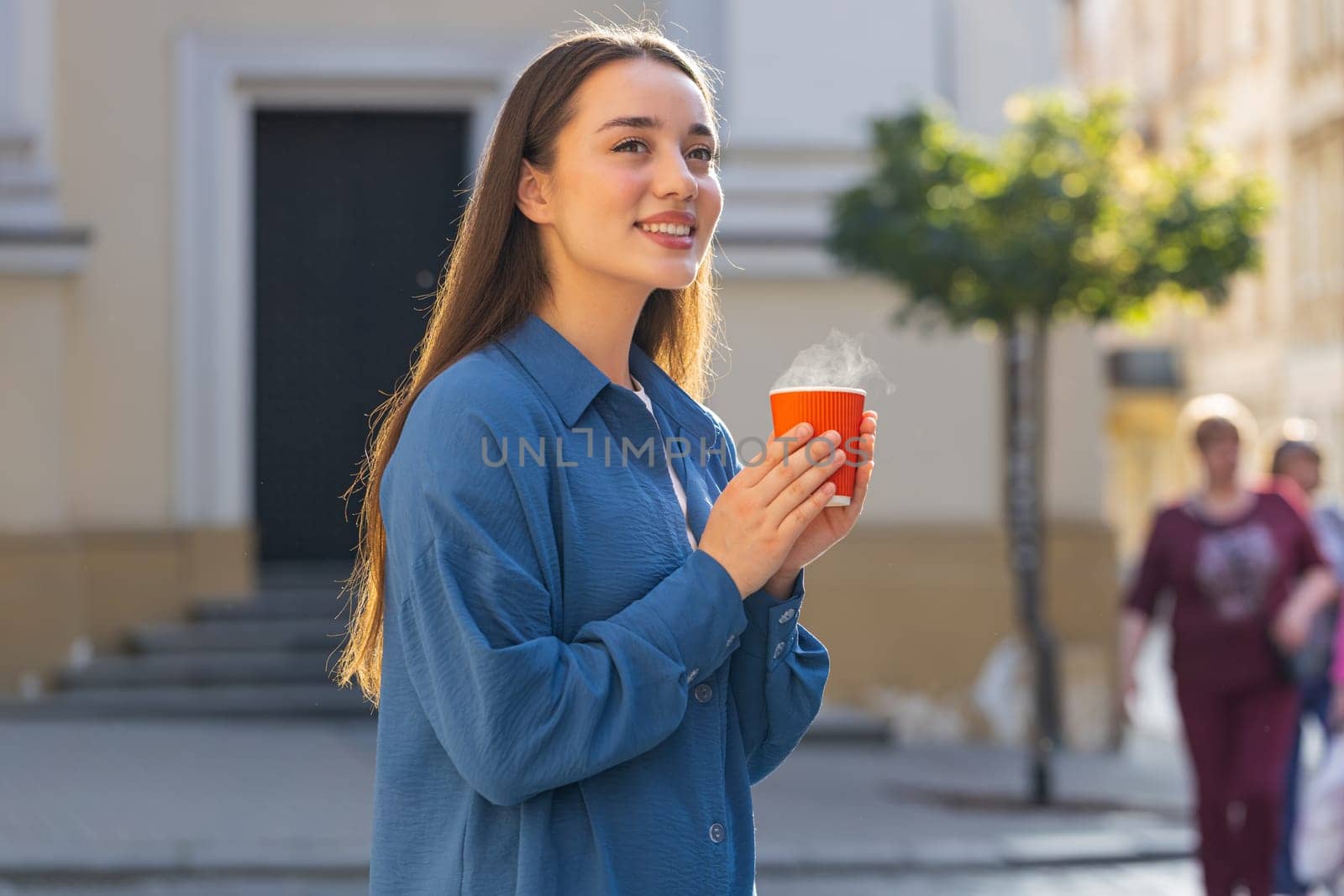 Portrait of pretty young woman enjoying morning aroma coffee steam or tea hot drink outdoors. Relaxing, taking a break. Lovely girl walking in urban city sunny street sun rays, drinking coffee to go