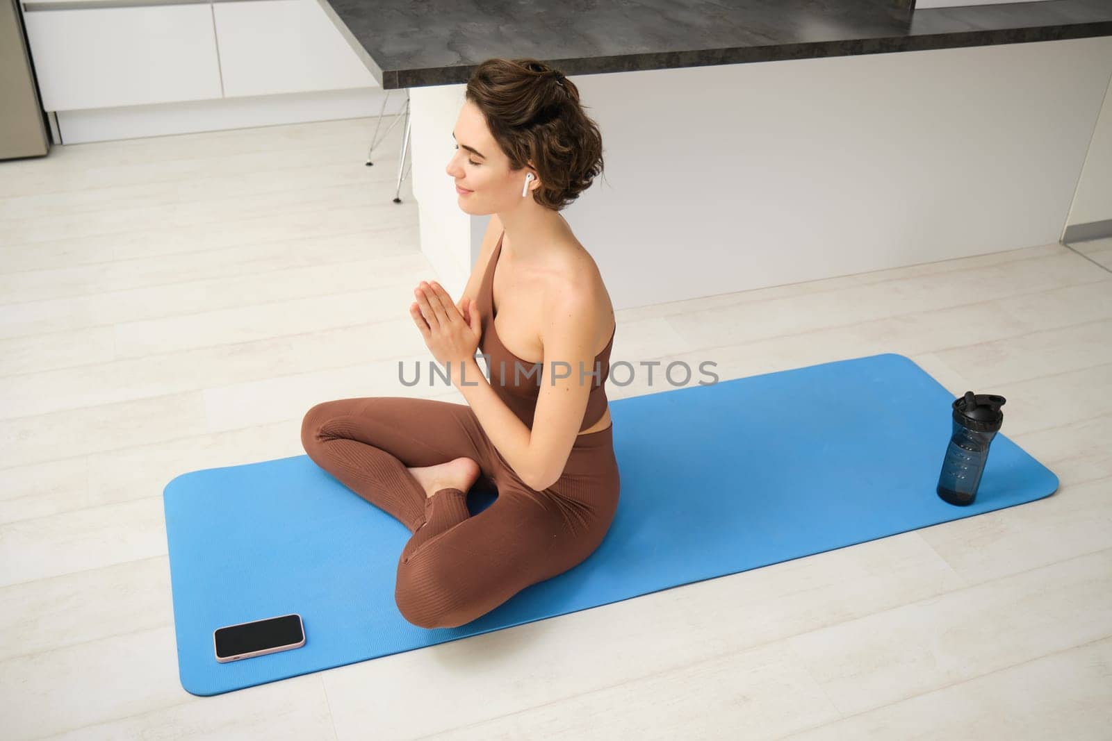 Young girl at home, sits in lotus pose, listens to meditation app in wireless headphones, smartphone and water bottle on her yoga mat, workout in kitchen, practice mindfulness.