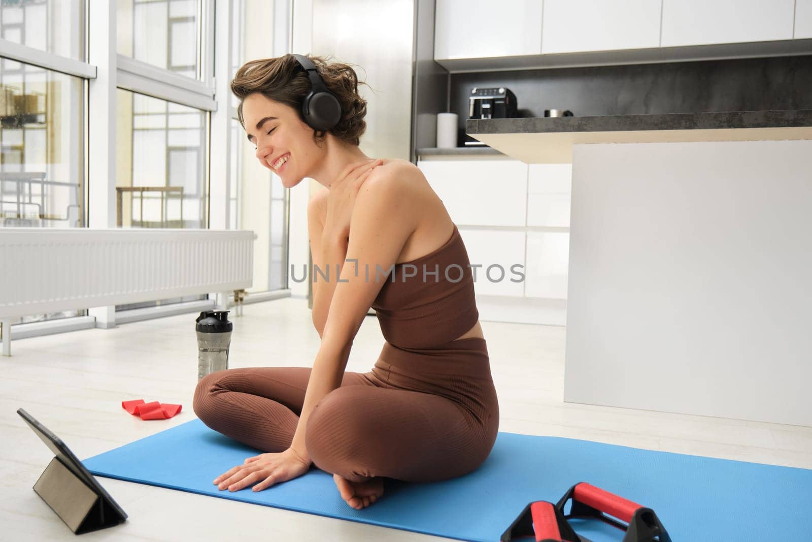 Smiling brunette fitness girl, listens music in headphones, watches online video from tablet, learns yoga poses, workout tips, does pilates exercises at home, indoor training and sport concept.