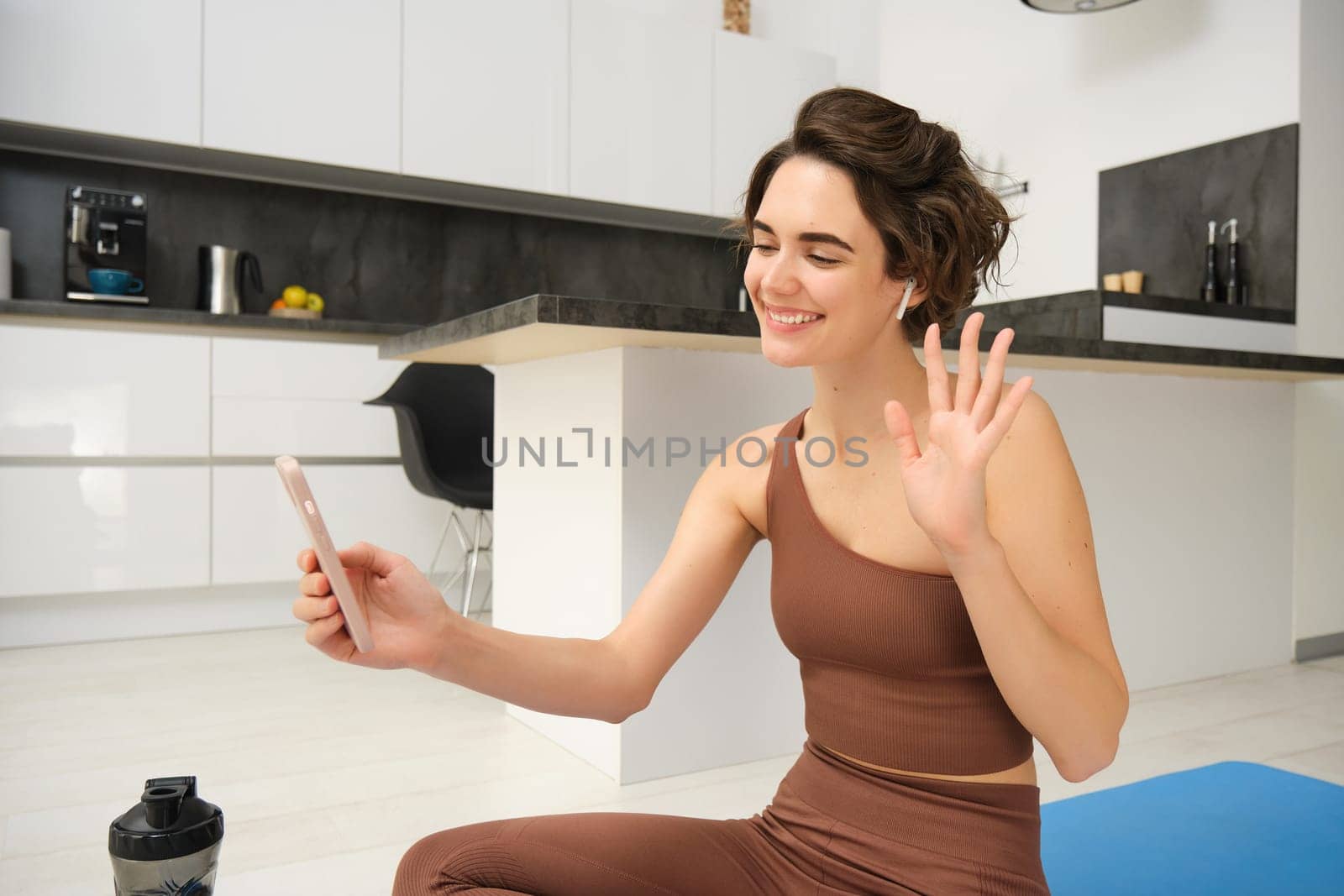 Cheerful young woman with mobile phone, waves and says hi at smartphone camera, connects to online gym class, remote yoga instructor with workout app, sits on yoga mat in kitchen at home.