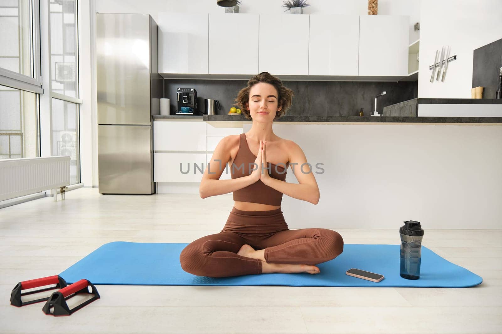 Smiling girl athlete, sportswoman doing yoga at home in activewear, sitting on yoga rubber mat in lotus pose, meditating, practice mindfulness exercises.