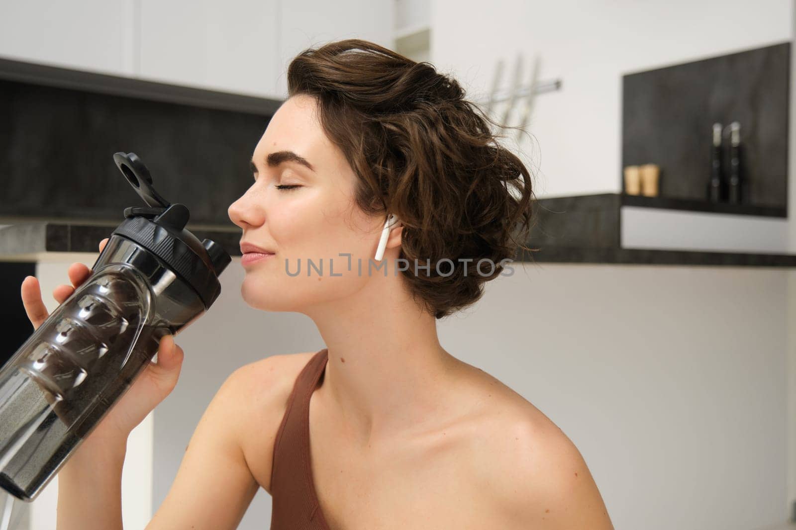 Portrait of fitness woman, young sportswoman at home, drinks water from bottle after workout, training exercises, takes a break after pilates, yoga training, stays hydrated.