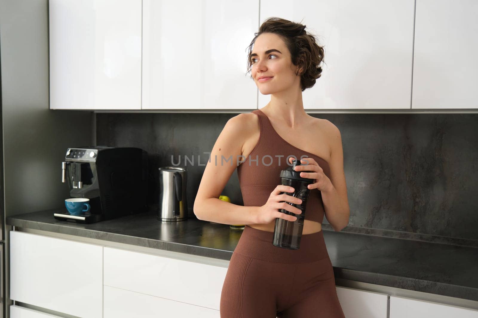 Sport and healthy lifestyle. Portrait of smiling fitness woman in activewear, standing near kitchen counter at home, drinking from water bottle, preparing for workout gym training.