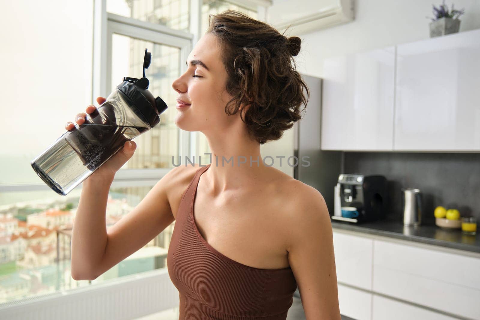 Healthy lifestyle and sport. Smiling brunette female athlete, fitness girl drinks water from gym bottle with pleased face, standing at home in kitchen.