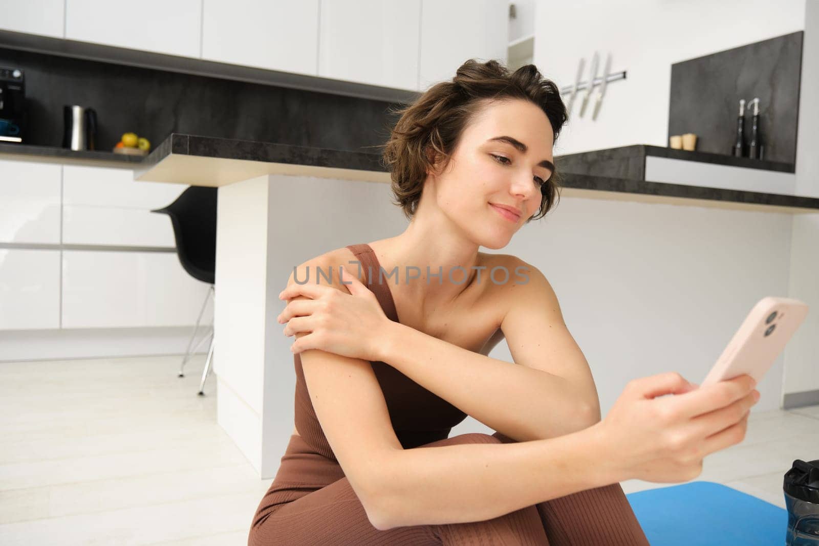 Sport and wellbeing. Portrait of fitness woman, gym instructor workout at home, sits on yoga mat and looks at her smartphone with smiling face.