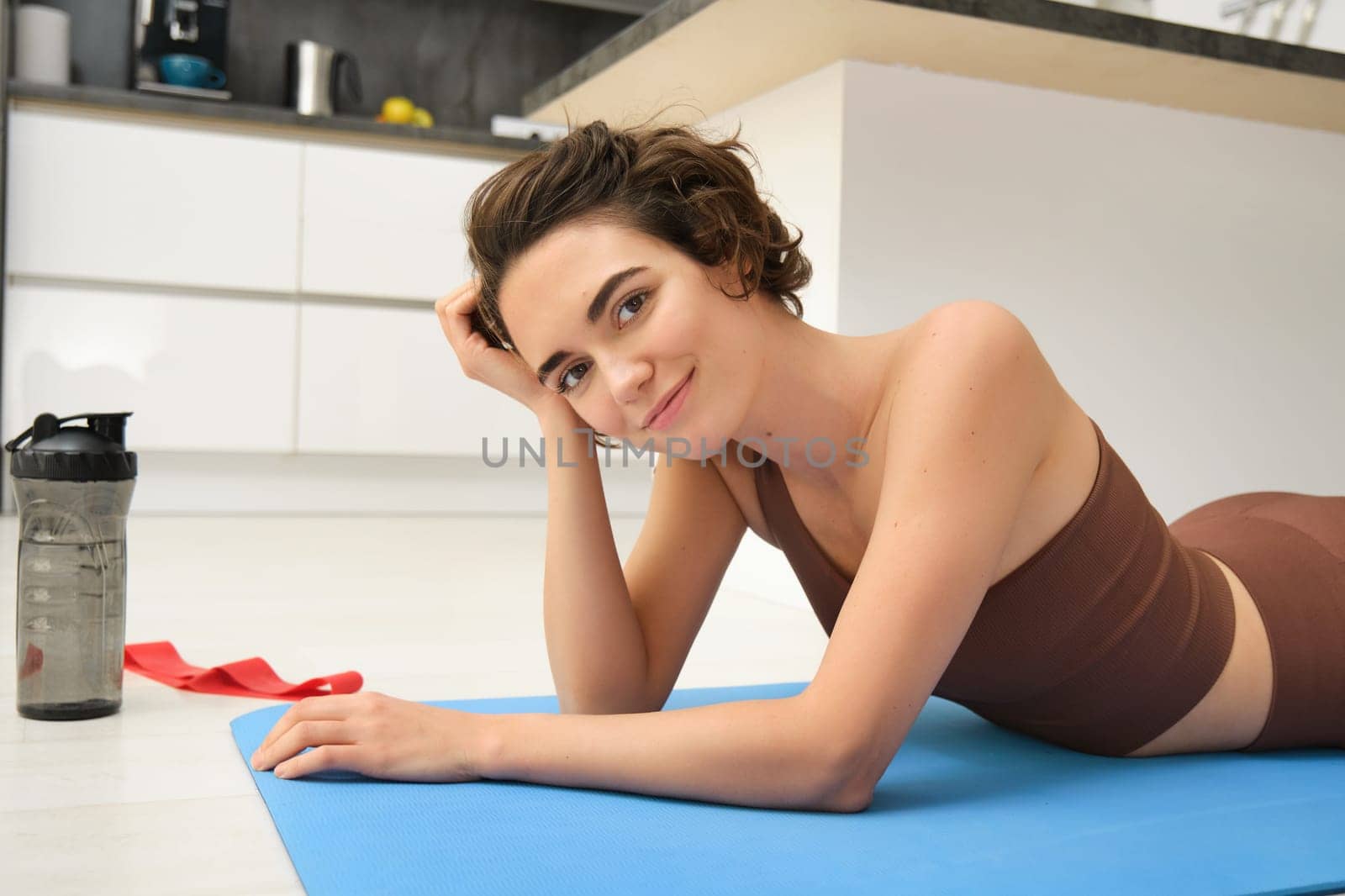 Portrait of fitness girl lying on rubber mat, warm-up before workout, doing pilates exercises at home, drinking water and doing sports, smiling at camera.