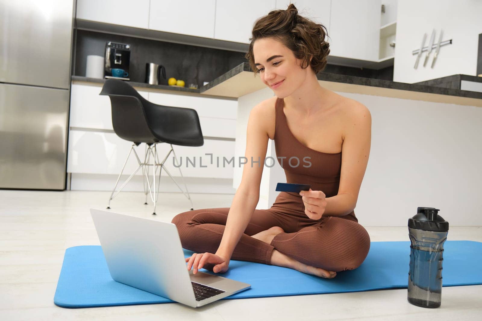 Portrait of young female athlete, yoga girl paying for online classes, purchasing remote training with gym instructor, sitting on her rubber mat with laptop and credit card.