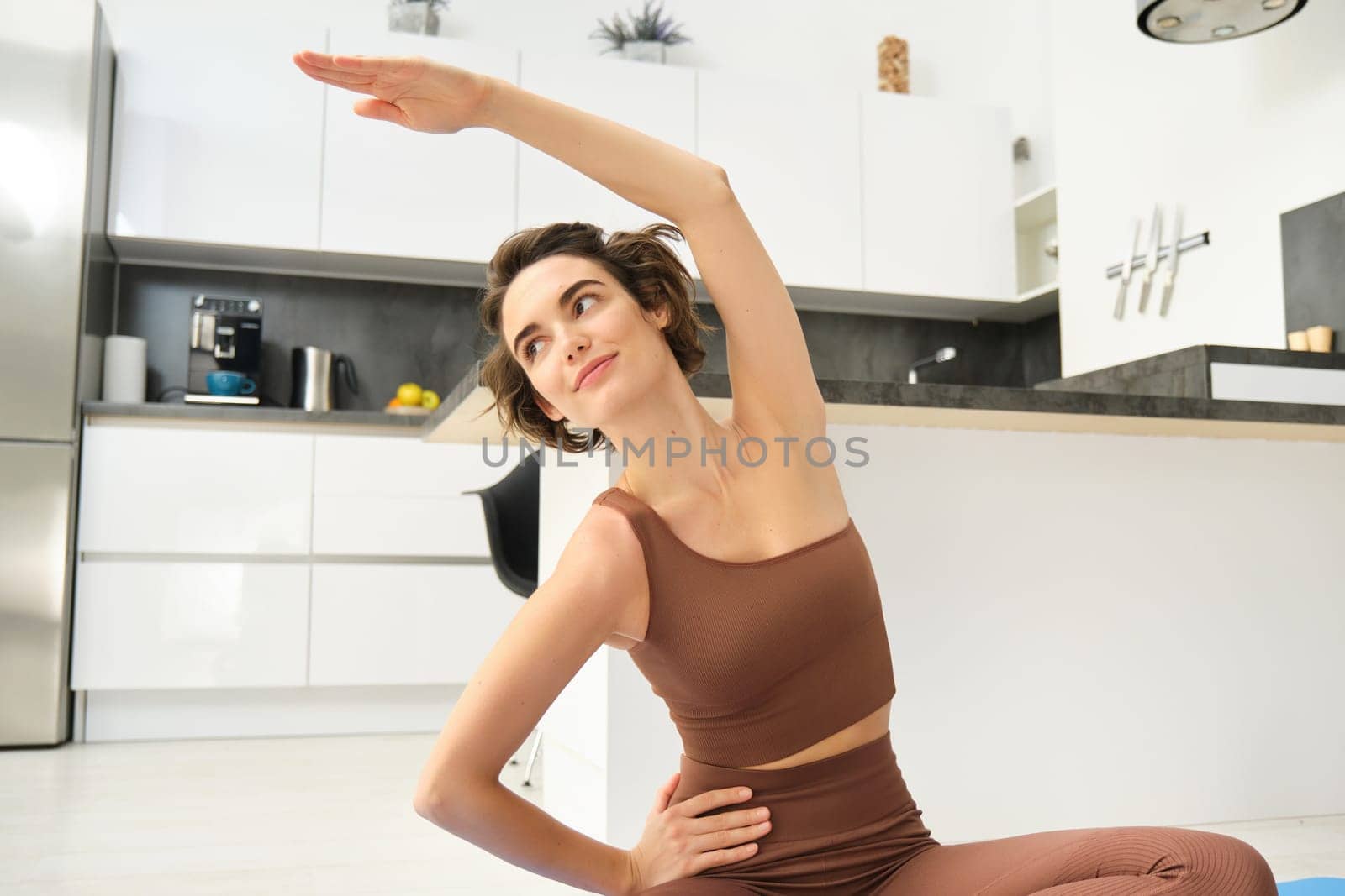 Fitness girl doing exercises at home. Young woman athlete sitting on yoga mat in bright room, stretching her arms, doing pilates exercises. Copy space