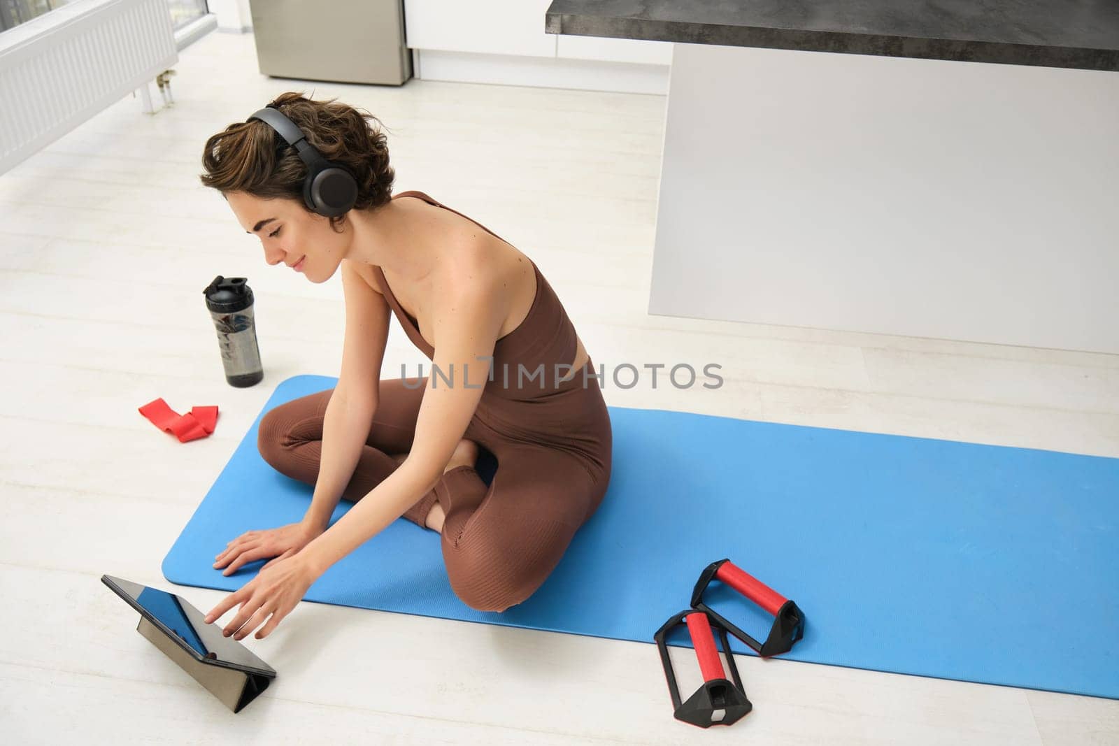 Smiling fitness girl in headphones, watches pilates, gym instructor online video, workout at home on yoga rubber mat, uses plank equipment and resistance band.