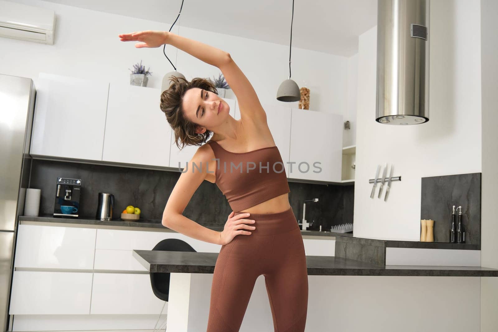 Home workout concept. Young woman in fitness clothes, doing stretching exercises, training at home behind kitchen, pulling arm to one side, focusing on body. Copy space
