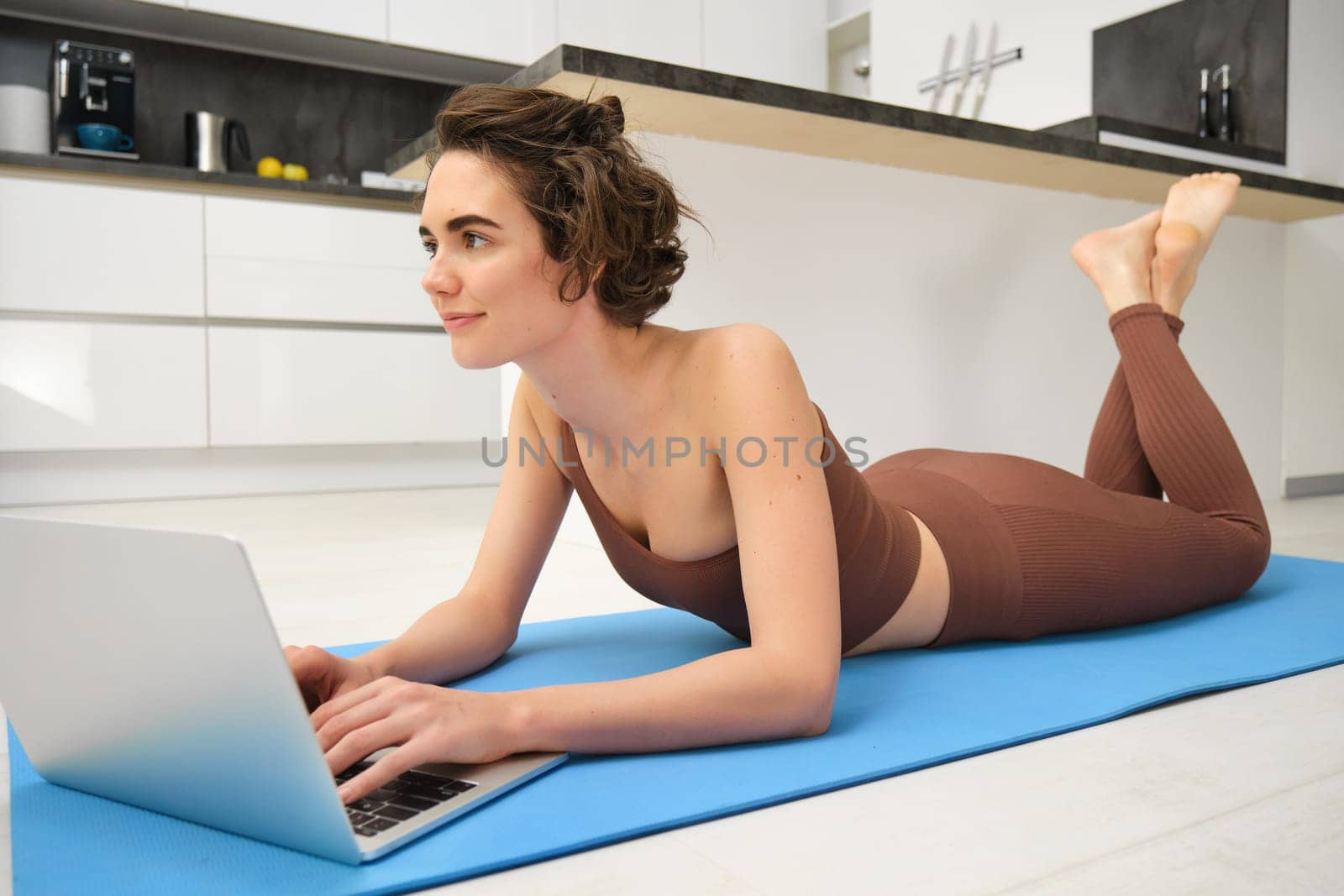 Home workout. Young fitness girl lying on yoga mat, using laptop in kitchen, training online with computer. Copy space