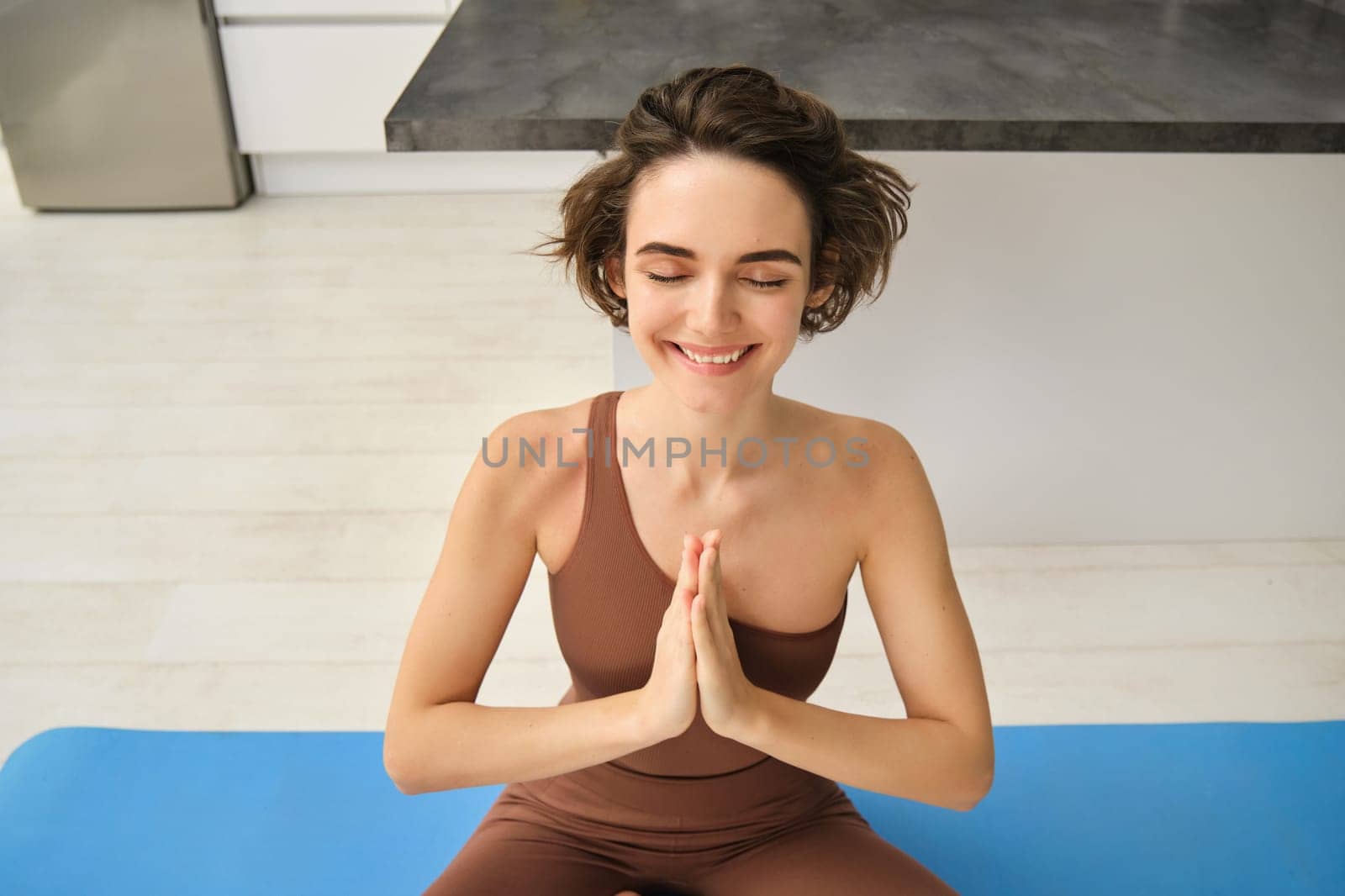 Peace, mindfulness and meditation. Young smiling sportswoman, fitness girl sits at home on floor and meditating, doing yoga, relaxing in lotus pose.