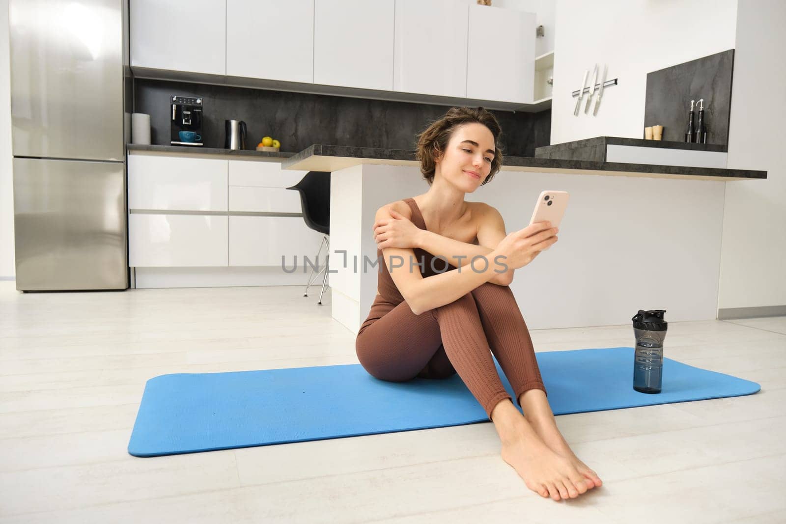 Portrait of sporty girl with mobile phone, sitting on yoga mat, using smartphone during workout at home, watching fitness turorial on telephone by Benzoix