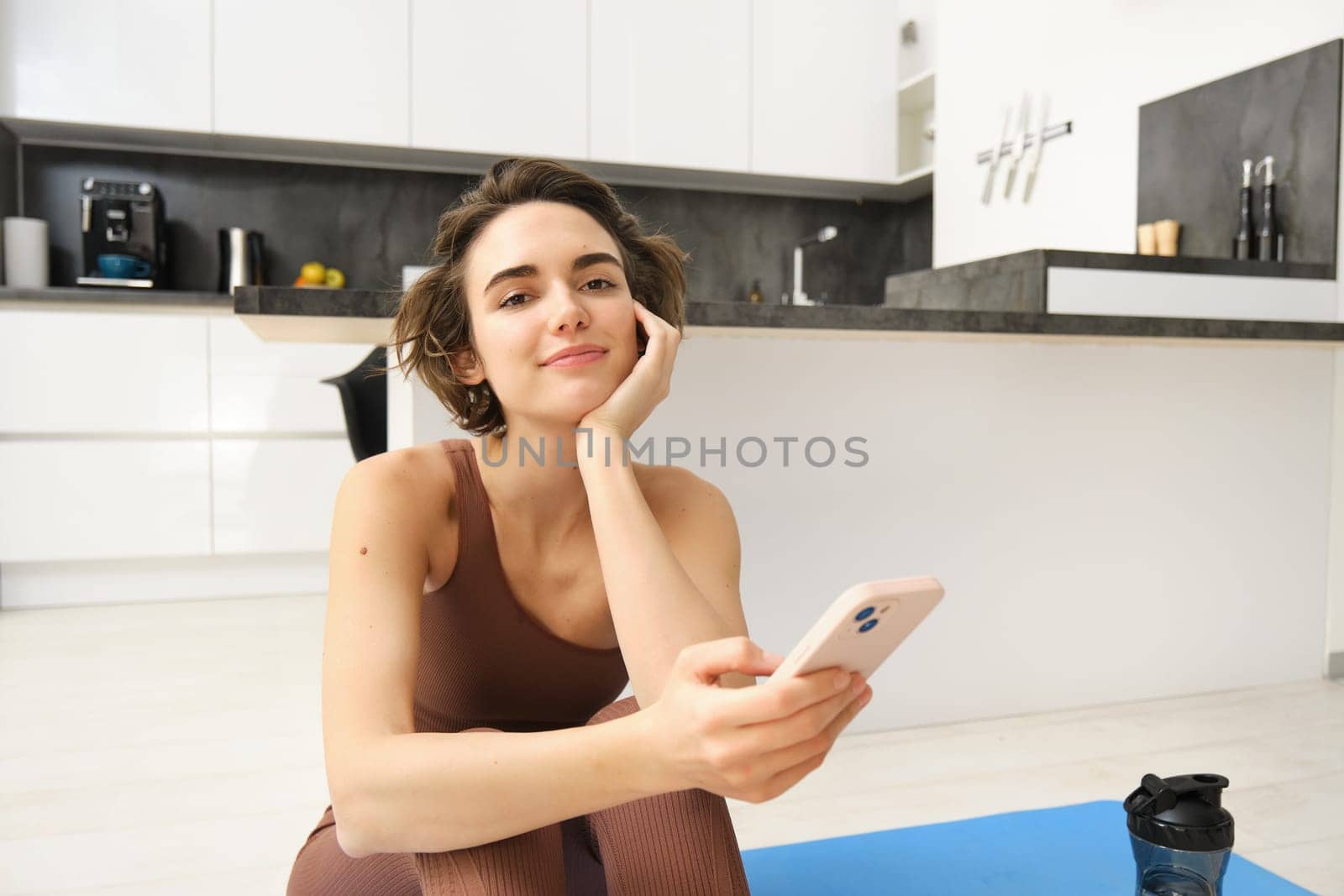 Technology and sport concept. Fitness girl looking at her smartphone, sitting in workout clothes at home, watching exercise videos on mobile phone by Benzoix