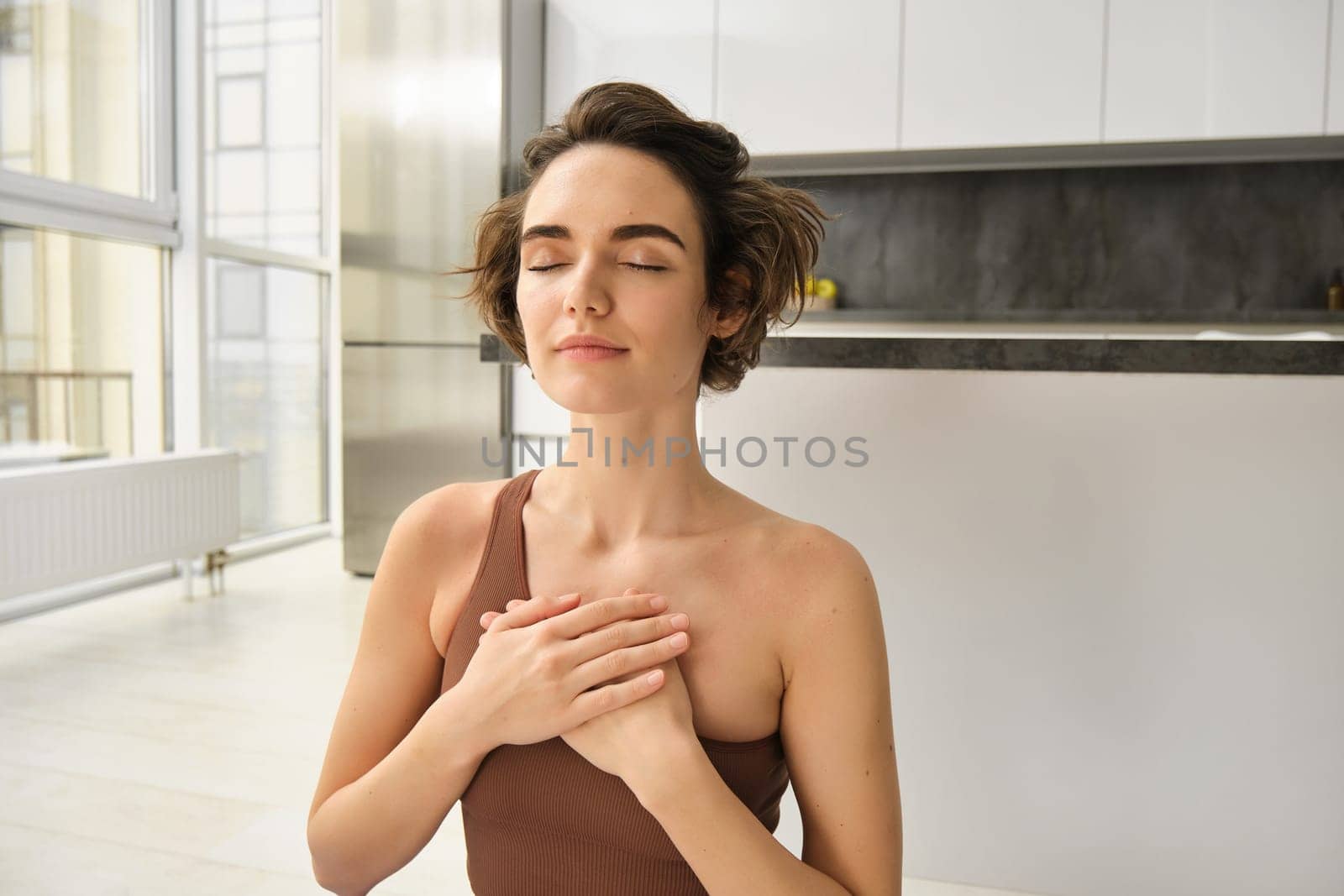 Portrait of young mindful woman, opens her chakra, doing yoga on rubber mat at home, closes eyes and smiles, feels peace inside her chest. Sport and lifestyle concept