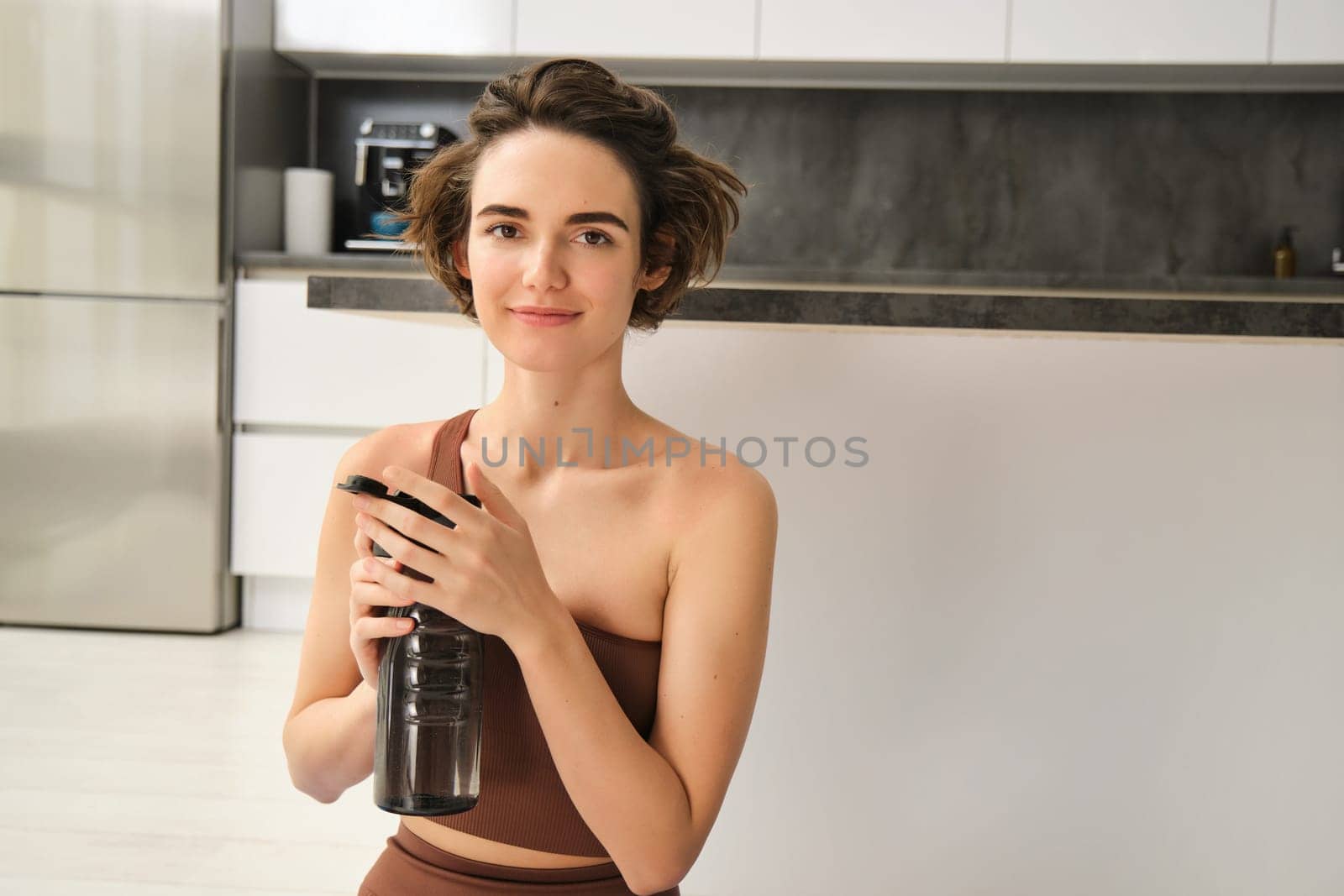 Fitness instructor, smiling young woman workout at home, sits on yoga mat and drinks water, does exercises, training session in her kitchen. Sport indoors concept
